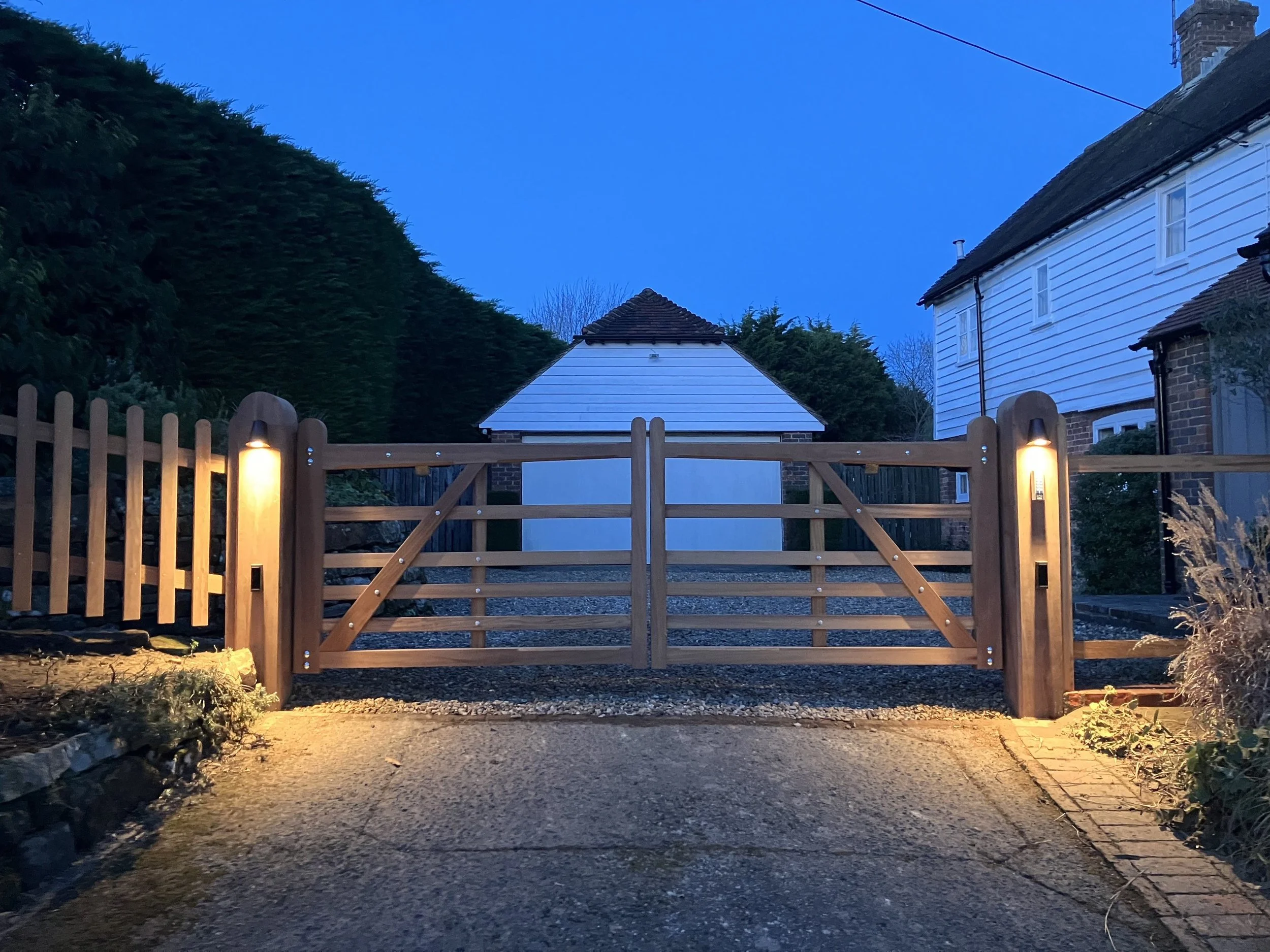 A wooden gate with lighting in front of a driveway, with houses and trees in the background during dusk or early evening.