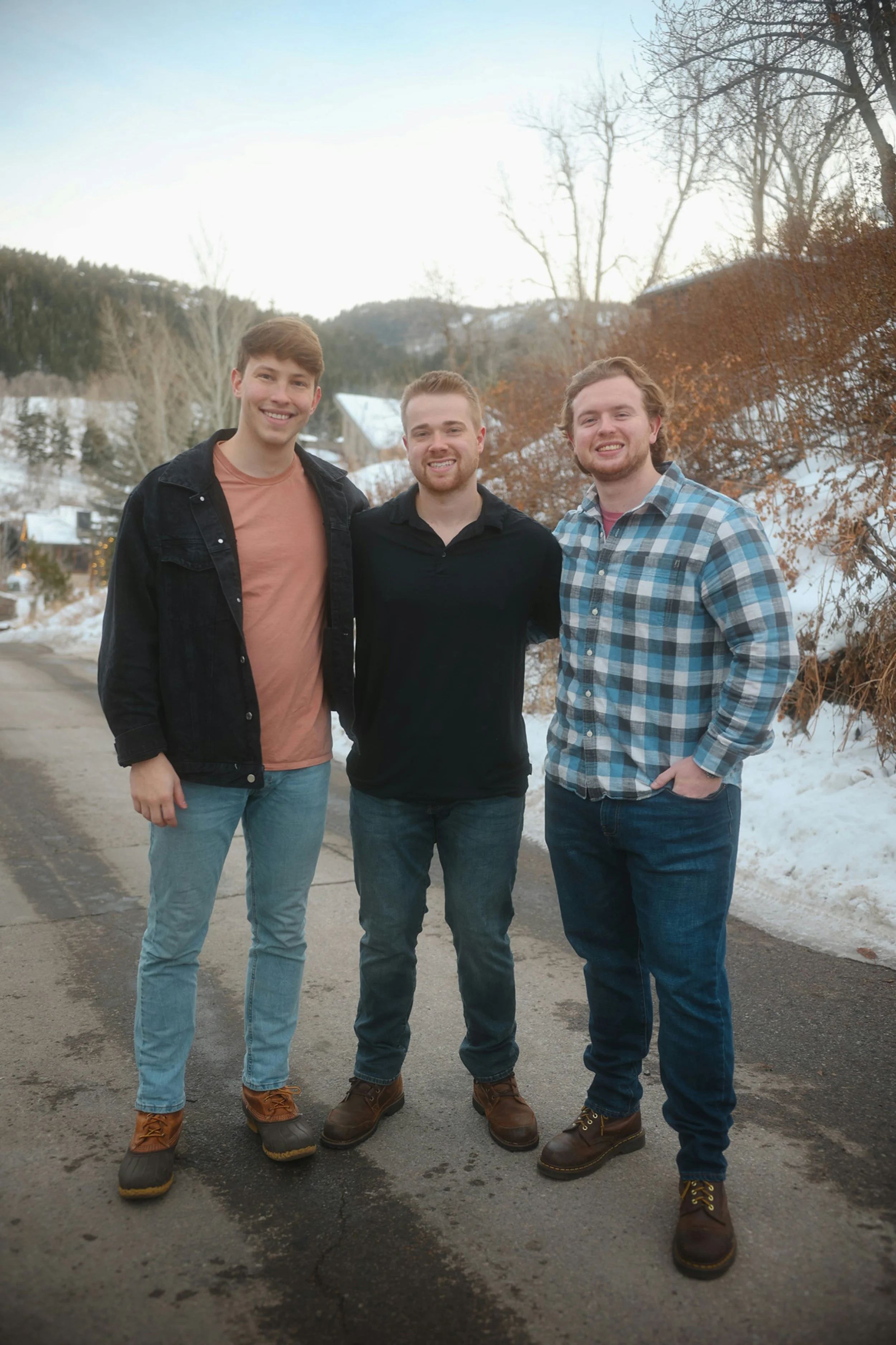 Three young men standing outdoors on a winter day, smiling at the camera. Snow on the ground and leafless trees in the background.