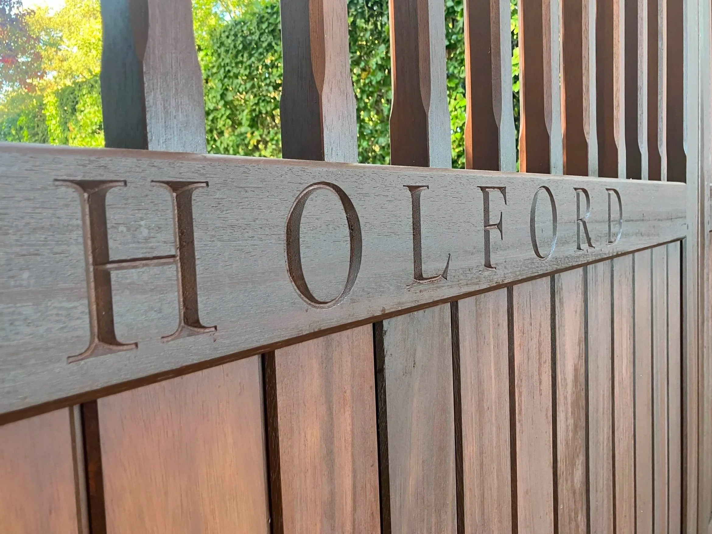 Wooden sign with engraved letters reading 'HOLD' mounted on a wooden fence with green foliage in the background.