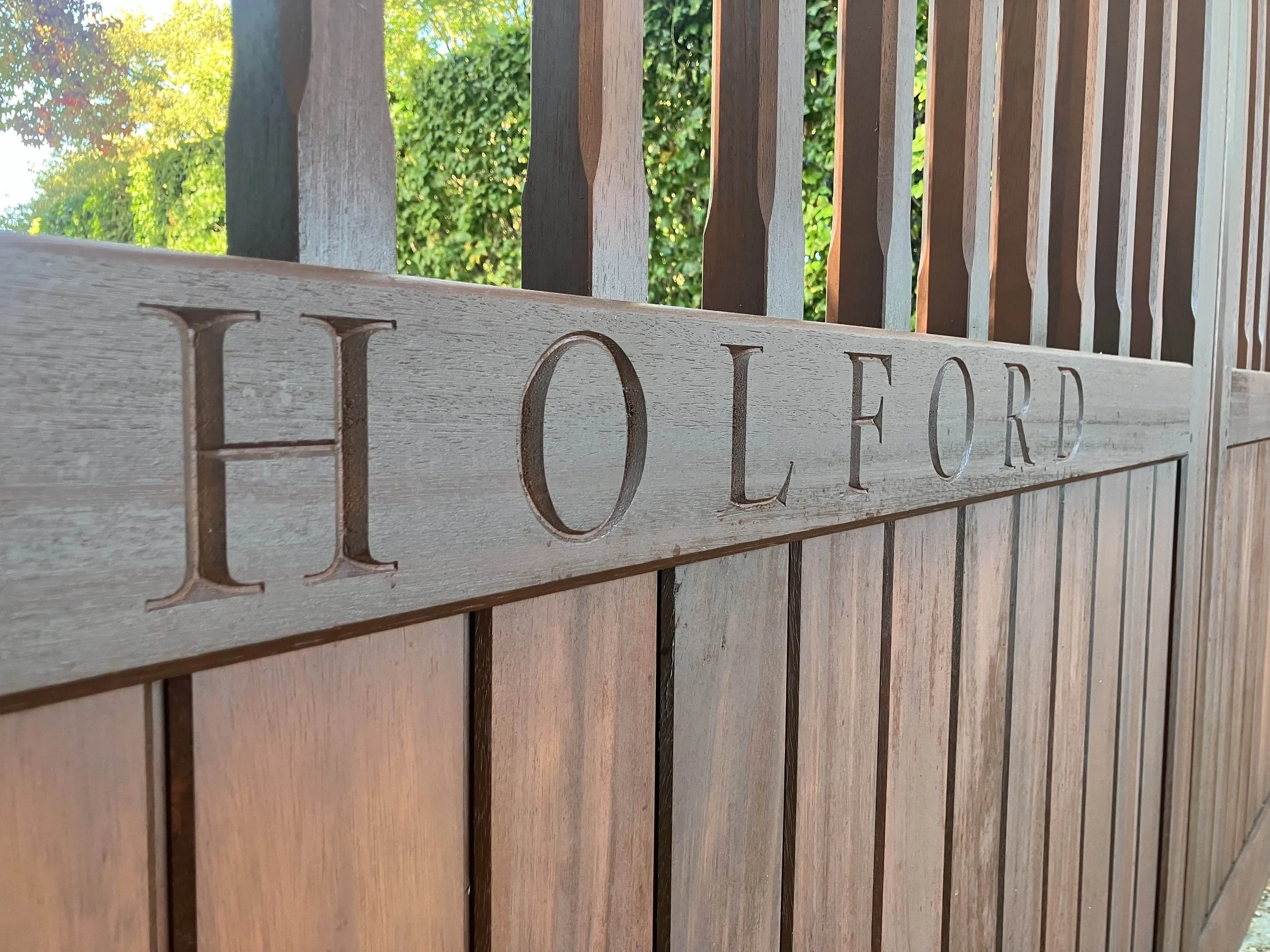 Wooden sign with partially visible word 'HOLD' behind a wooden fence with vertical slats. Green trees and sunlight in the background.