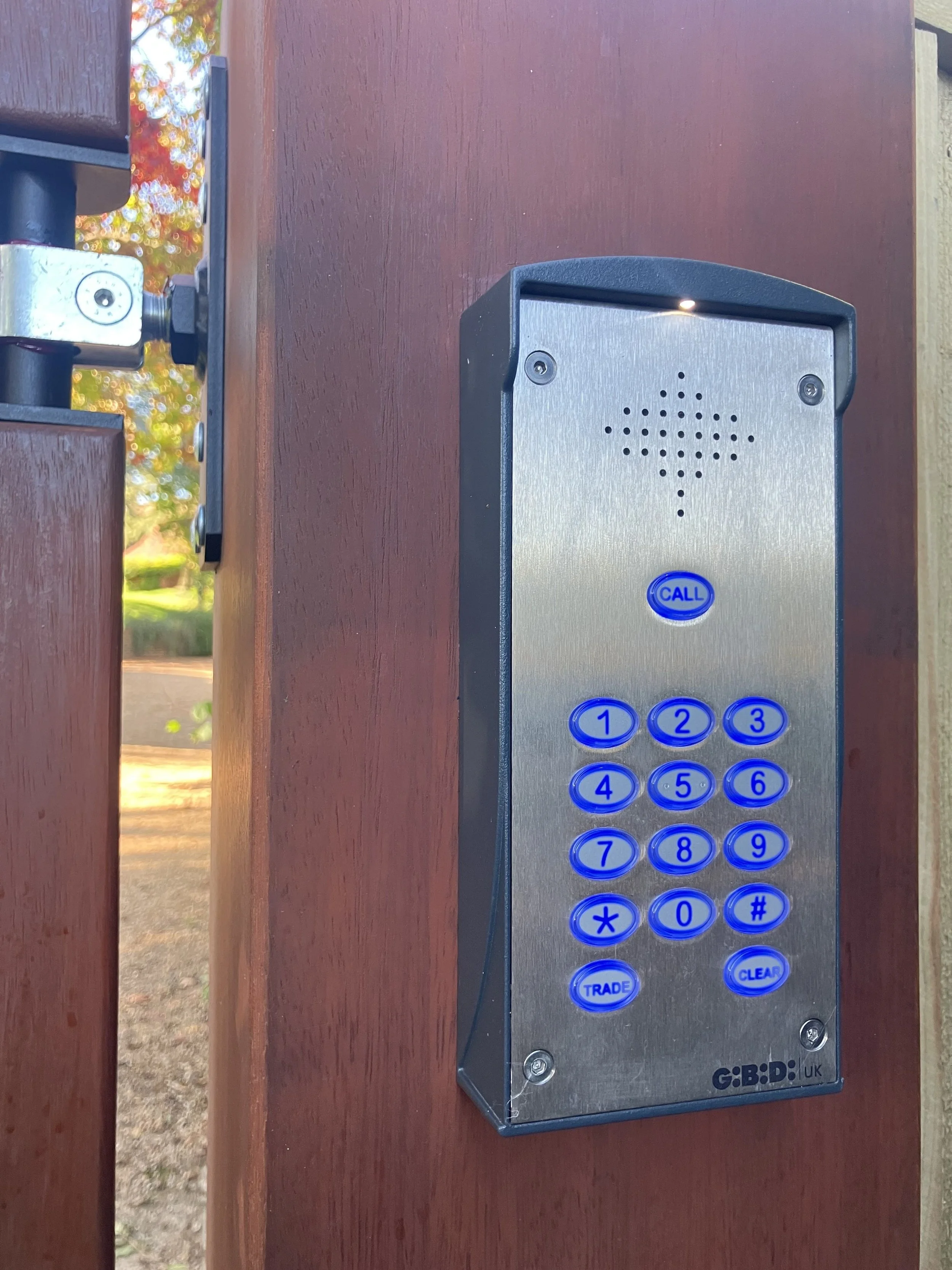 A metallic intercom with numbered blue buttons and a call button mounted on a wooden gate, with trees and sunlight visible in the background.