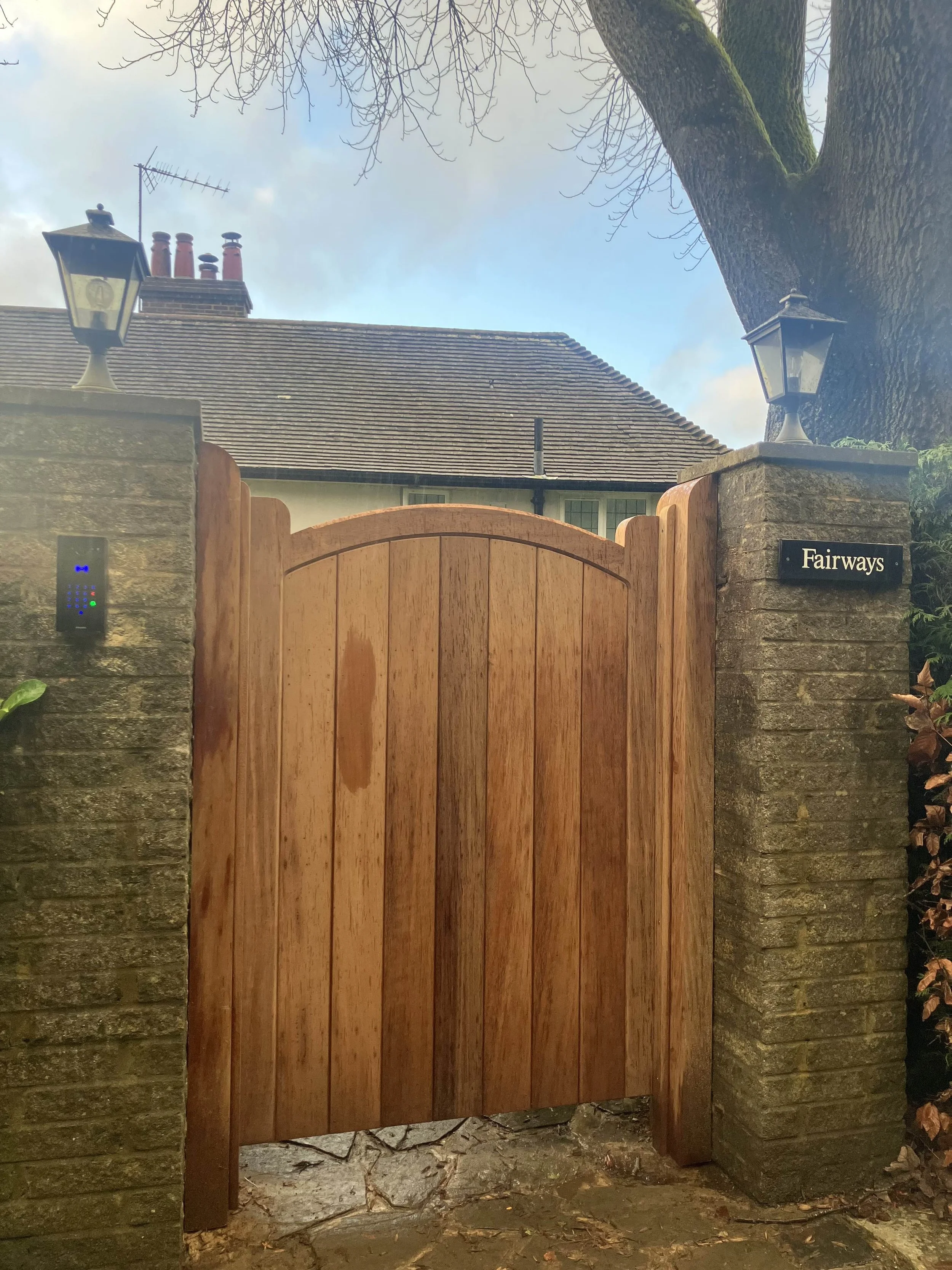 A wooden gate framed by stone pillars with lanterns on top, a stone wall on the side, a large tree with bare branches, and a house with a tiled roof in the background. The sign on the right stone pillar reads 'Fairways'.
