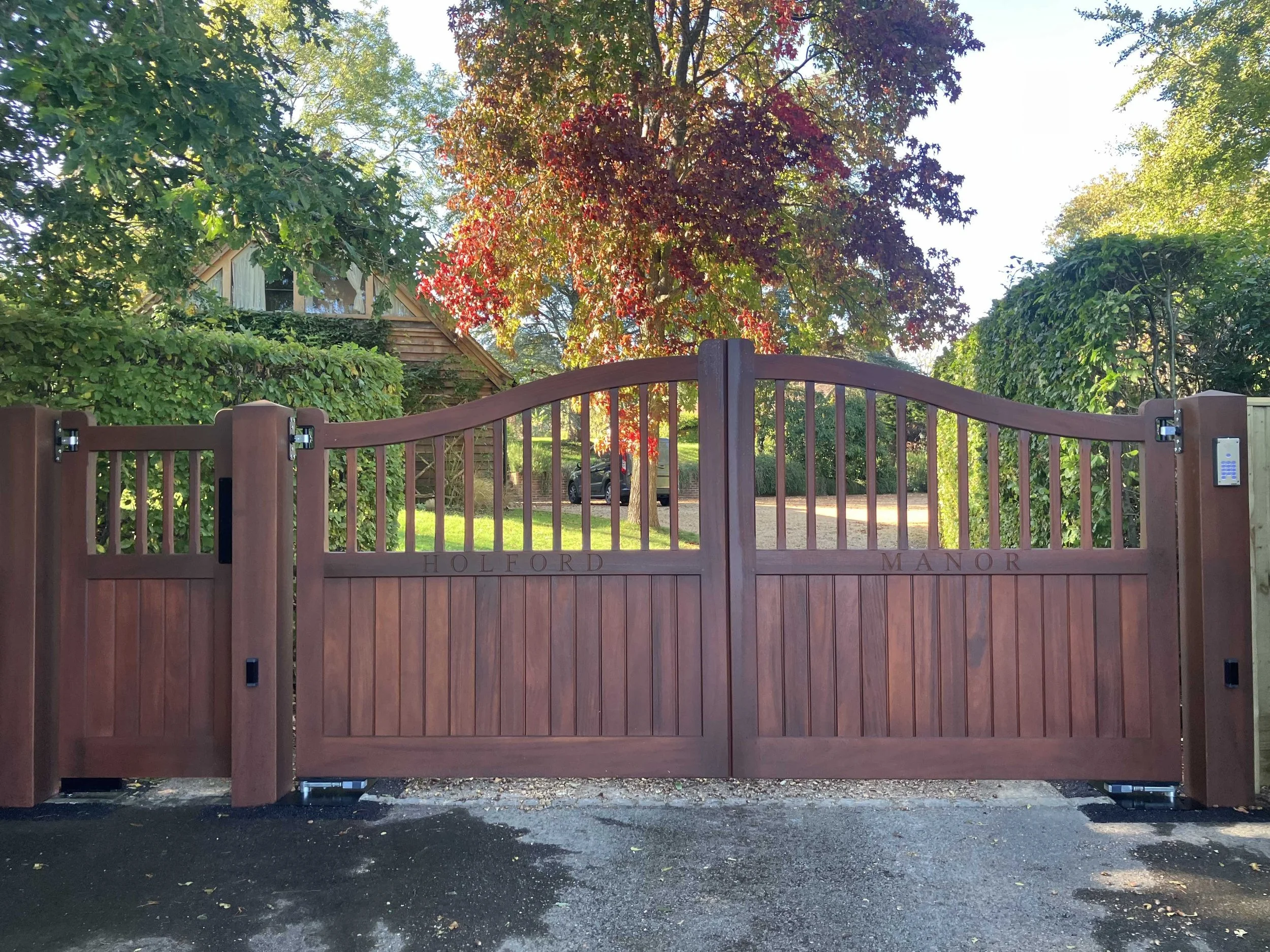 A large, brown wooden gate with the engraved words 'HOLFORD MANOR' at the bottom. Behind the gate, there is a tree with red and green leaves, a house, and a parked car in a lush green yard. The scene is illuminated by sunlight, casting shadows on the