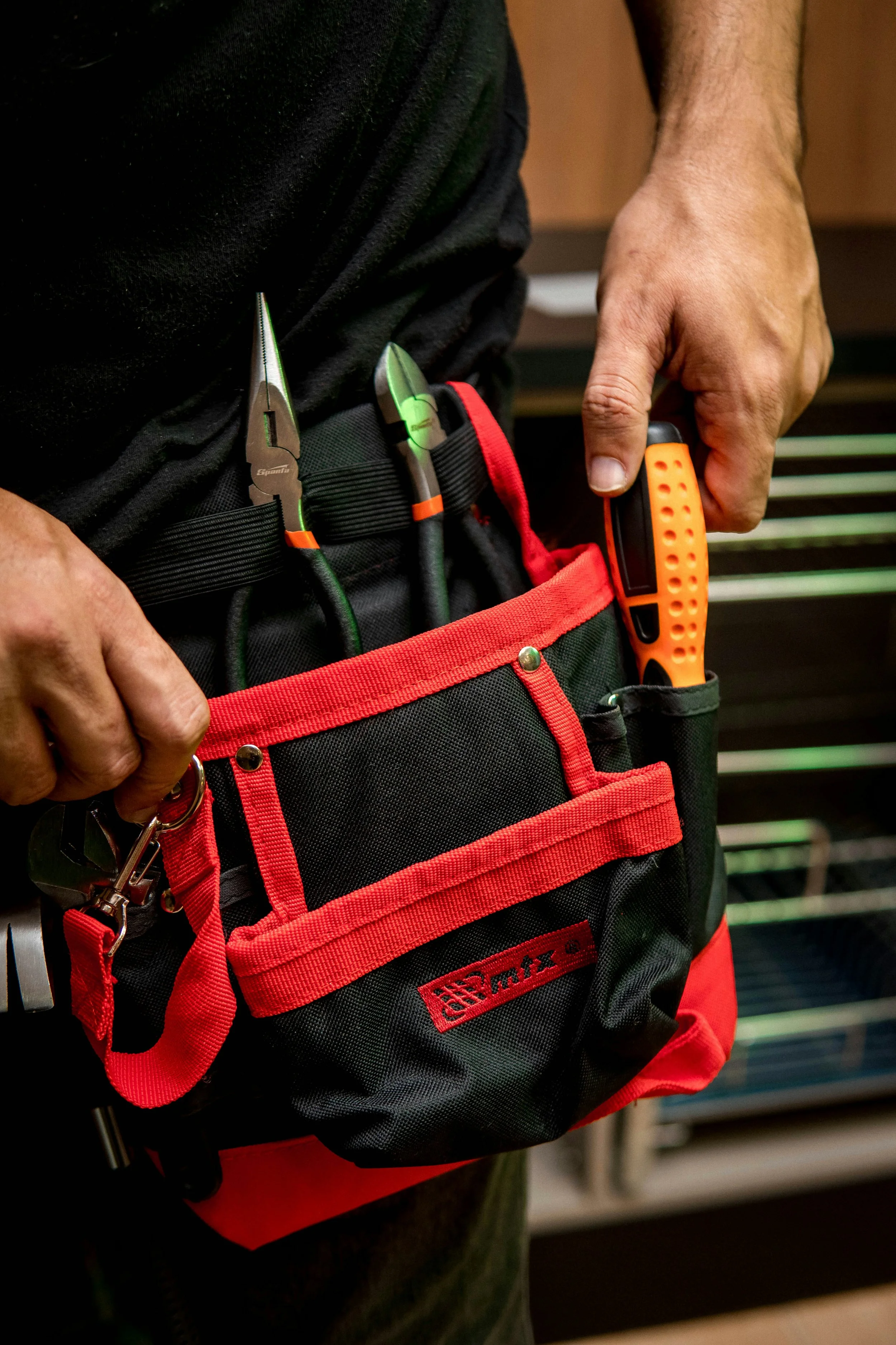 Close-up of a person wearing a tool belt with three screwdrivers and other tools, standing in front of a blurred background.