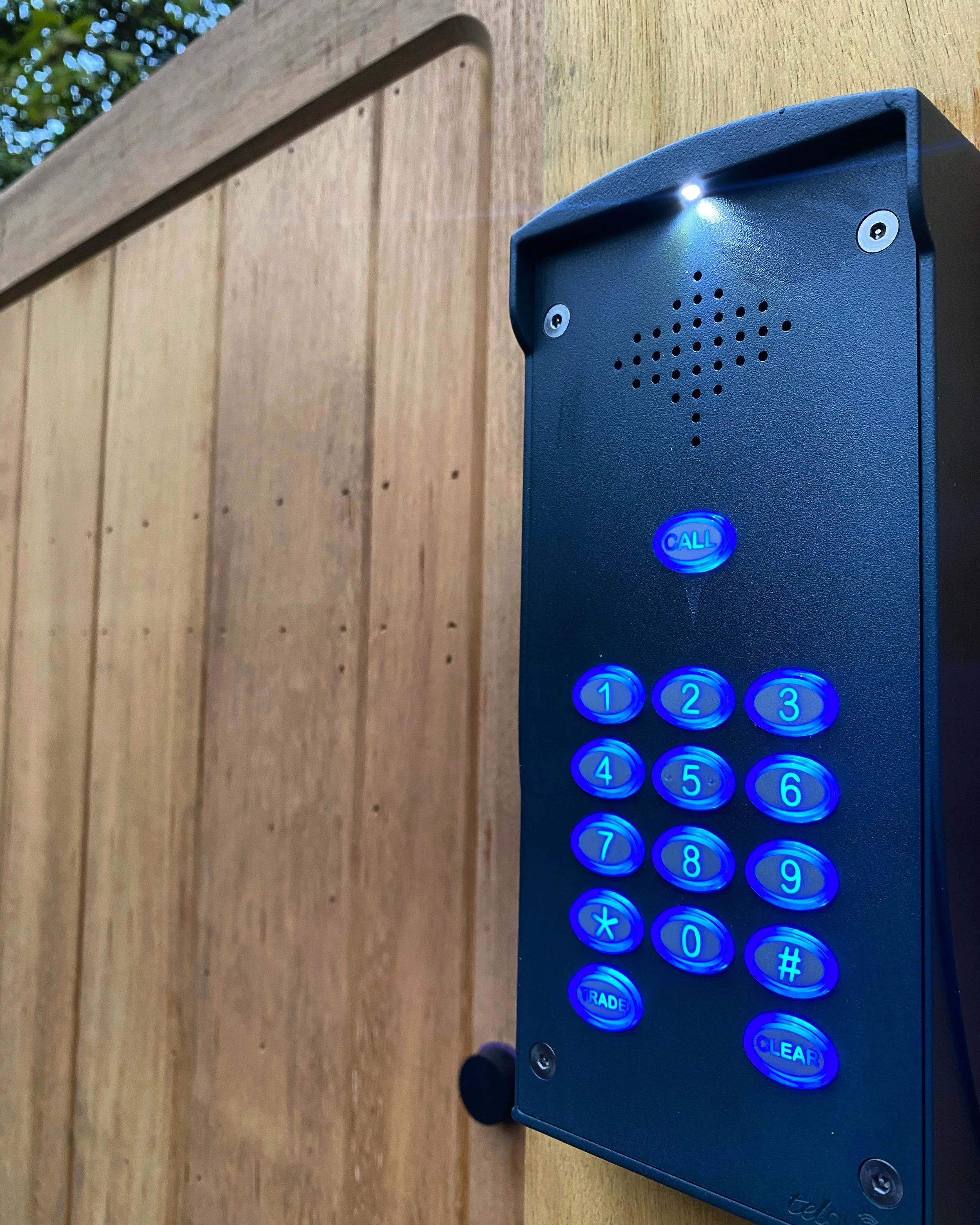 A blue outdoor intercom with illuminated call button and numeric keypad mounted on a wooden post.