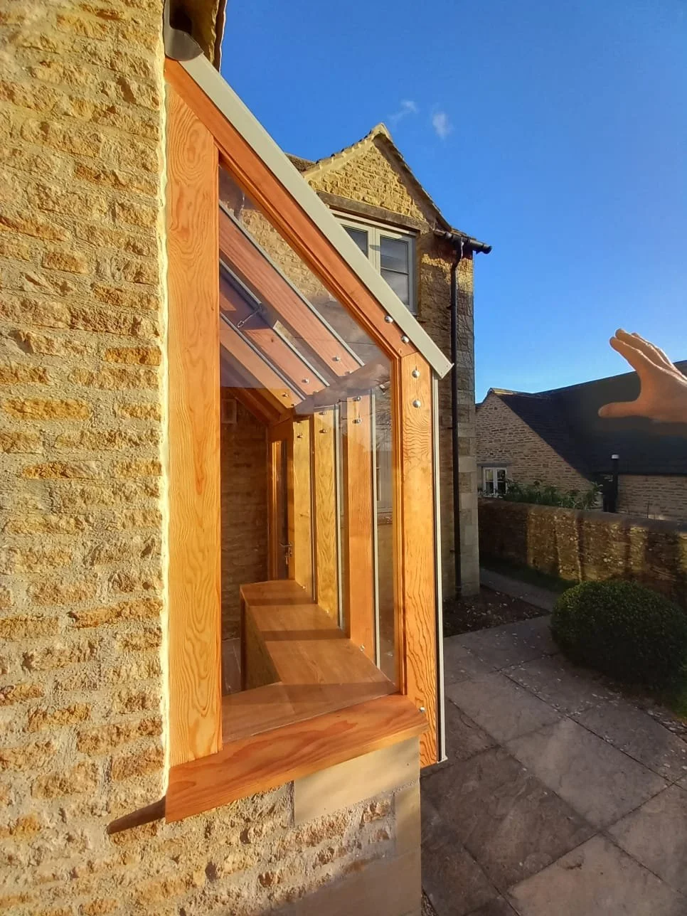 A photo of a small outdoor wooden and glass structure attached to a brick house, with a clear blue sky and neighboring houses visible in the background.