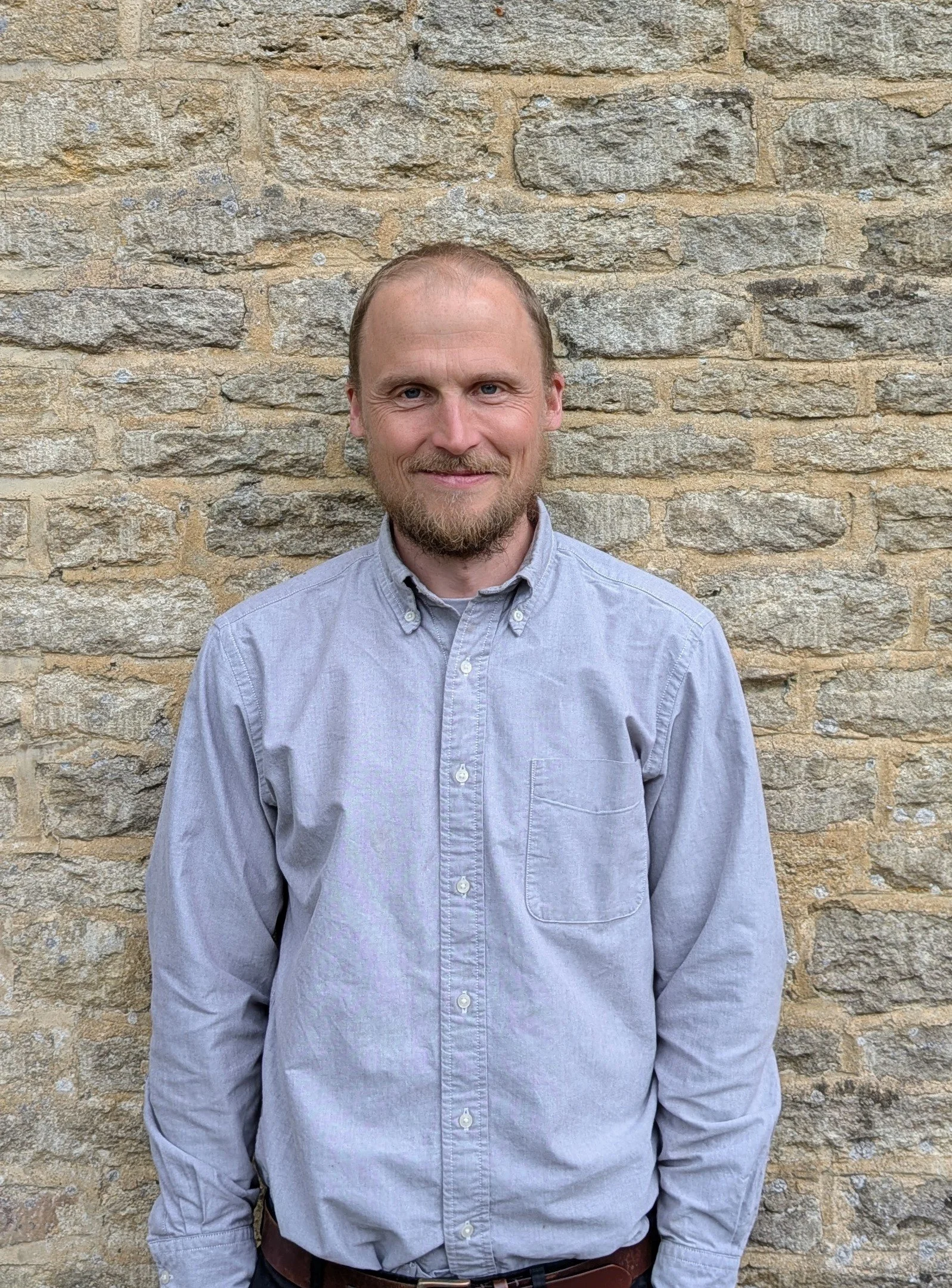 A man with a beard and light hair, wearing a light gray button-up shirt, standing in front of a beige brick wall.