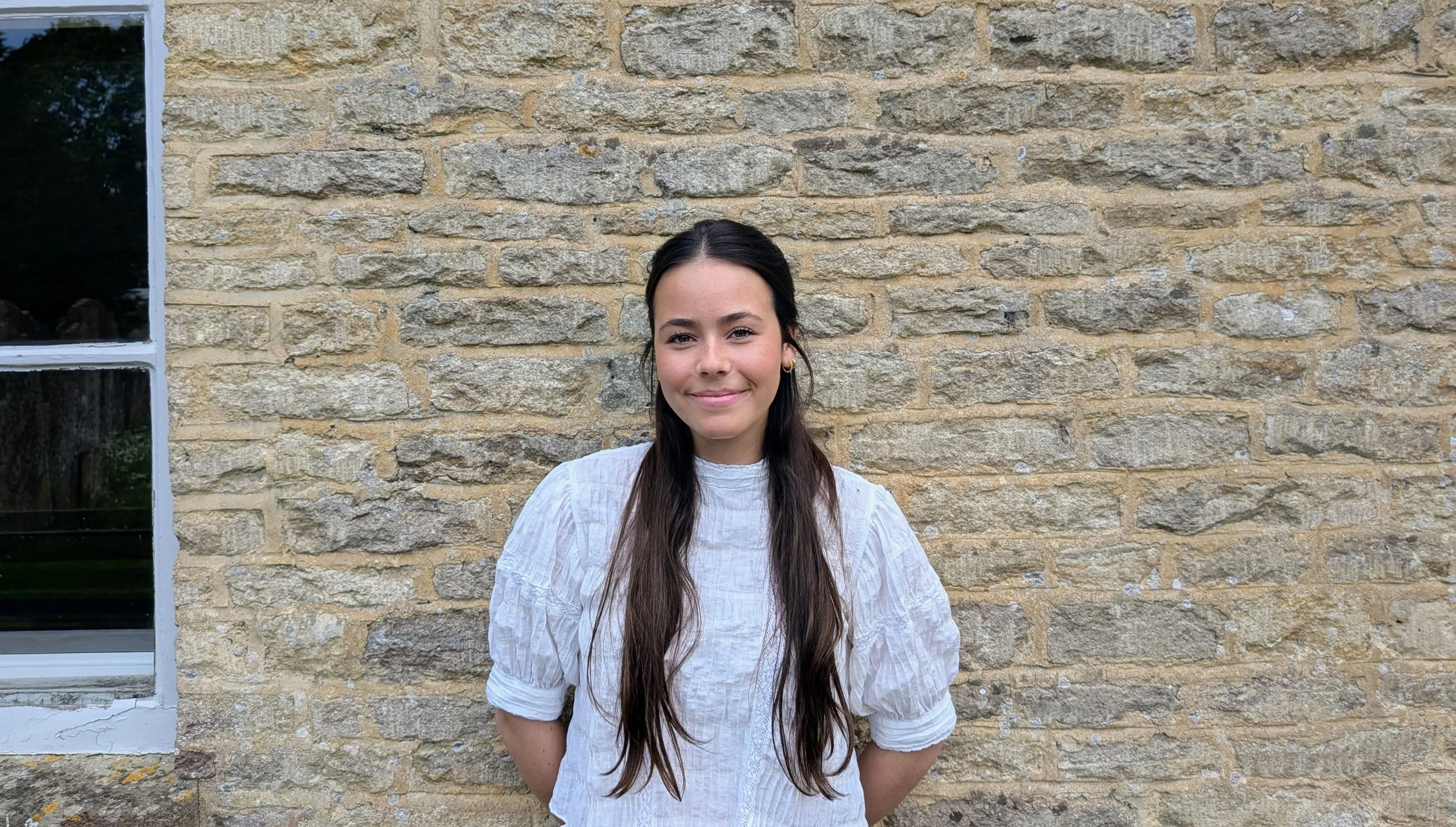 Young woman with long dark hair, wearing a white blouse, standing in front of a beige stone brick wall with a window on the left.
