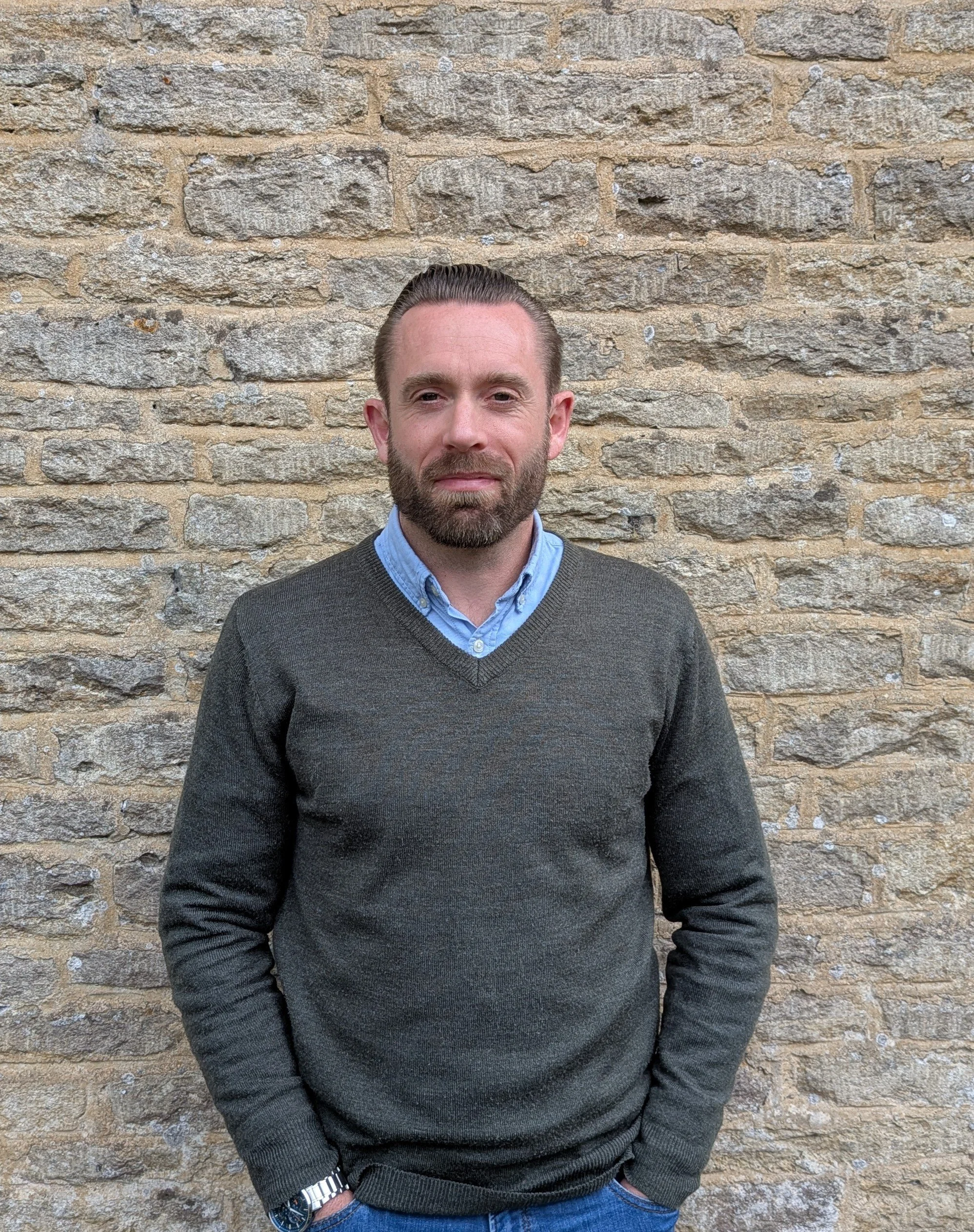 A man with a beard and slicked-back hair wearing a blue collared shirt under a dark sweater, standing against a beige brick wall.