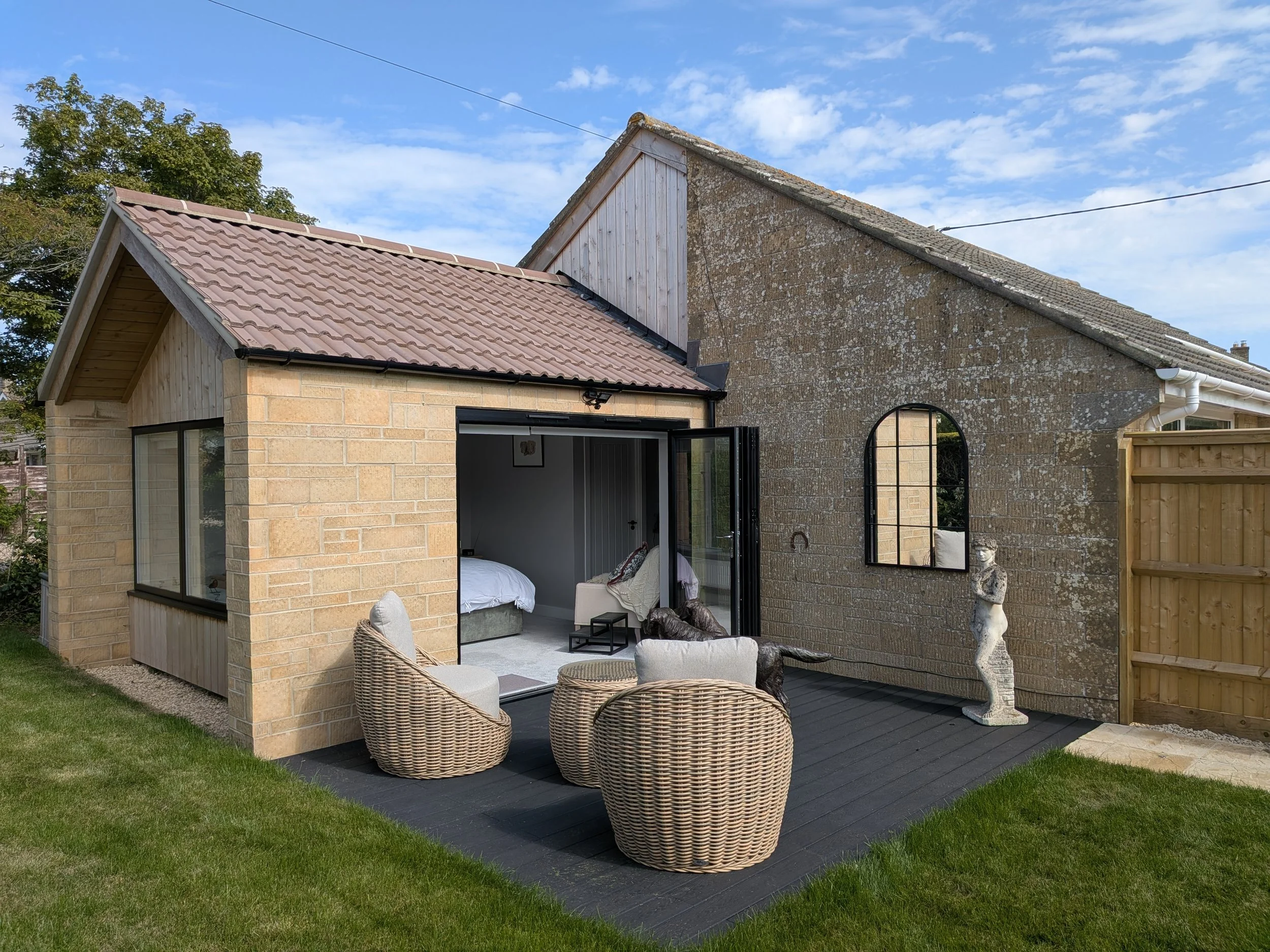 Backyard patio area with outdoor furniture, wicker chairs, and a white dog statue next to a wooden fence, with a house featuring a brick exterior and a mix of new and weathered roofing, under a blue sky with clouds.