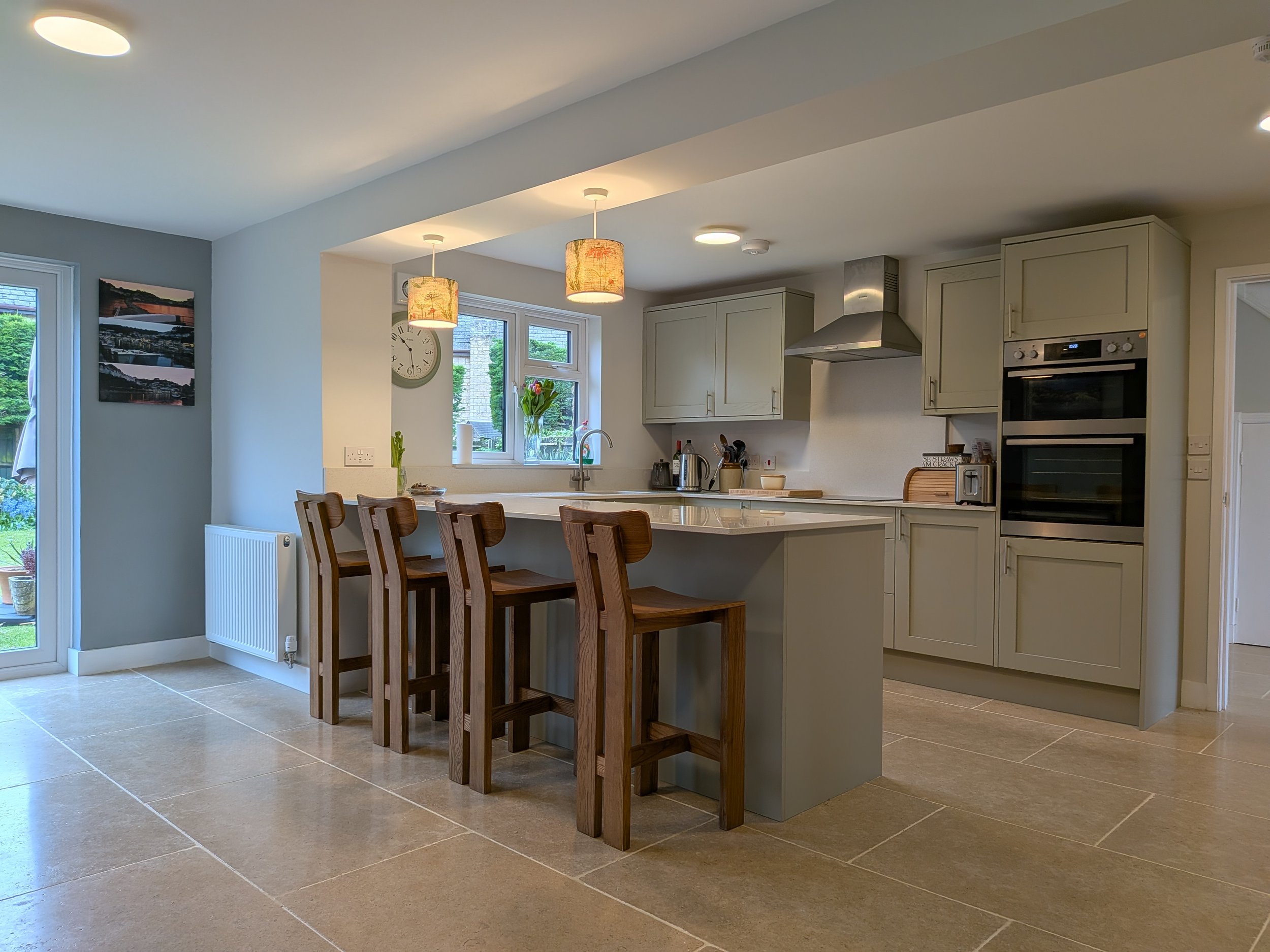 A modern kitchen with white cabinets, a countertop with a flower vase, a window, and a set of four wooden chairs at the kitchen island. There is a double oven and a stainless steel range hood.