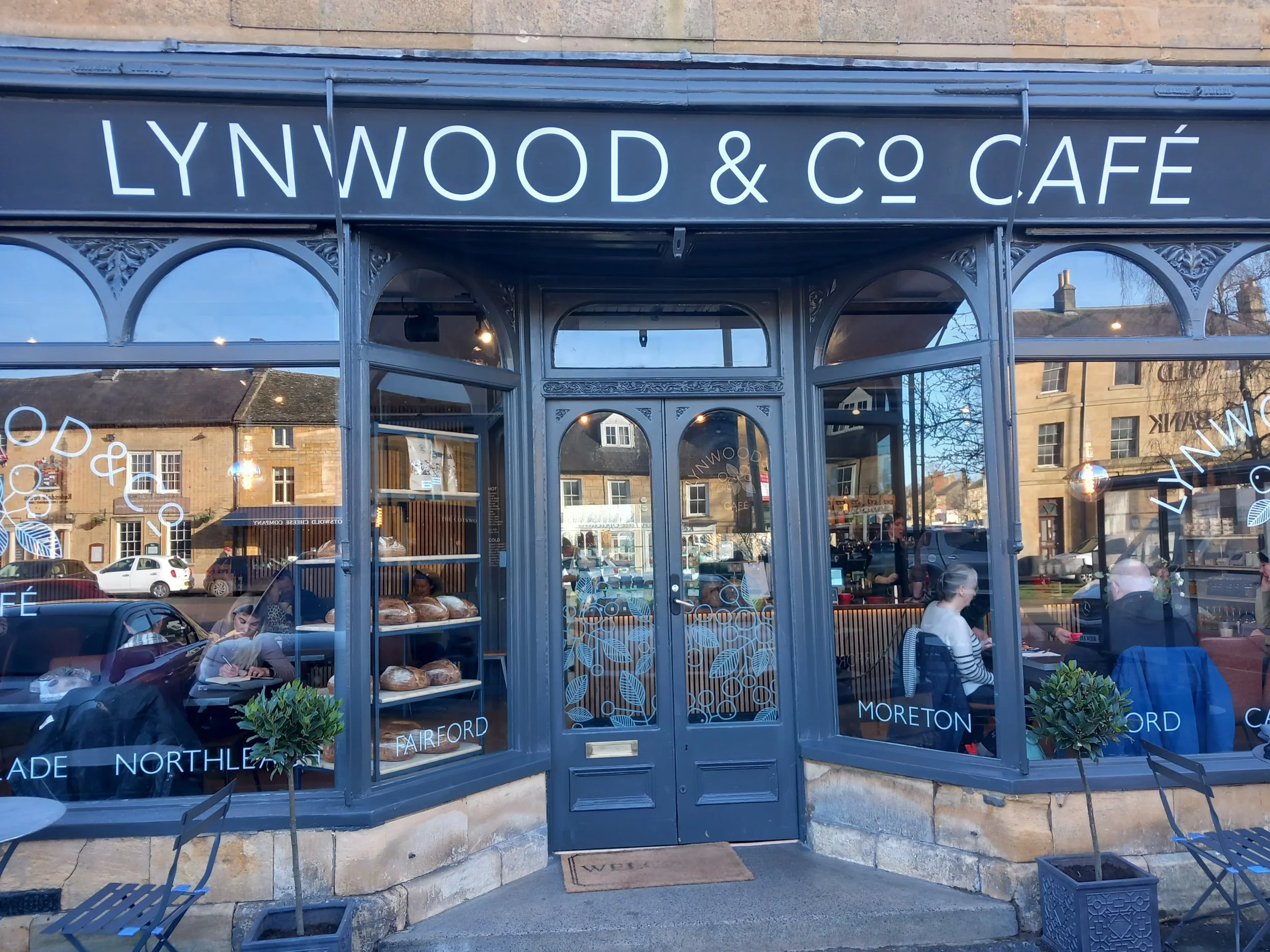 The exterior of Lynwood & Co Café with large glass windows and a black sign with white lettering. Inside, baked goods are visible through the window, and people are dining. The café is located in a historic building with stone walls and additional buildings can be seen reflected in the glass.
