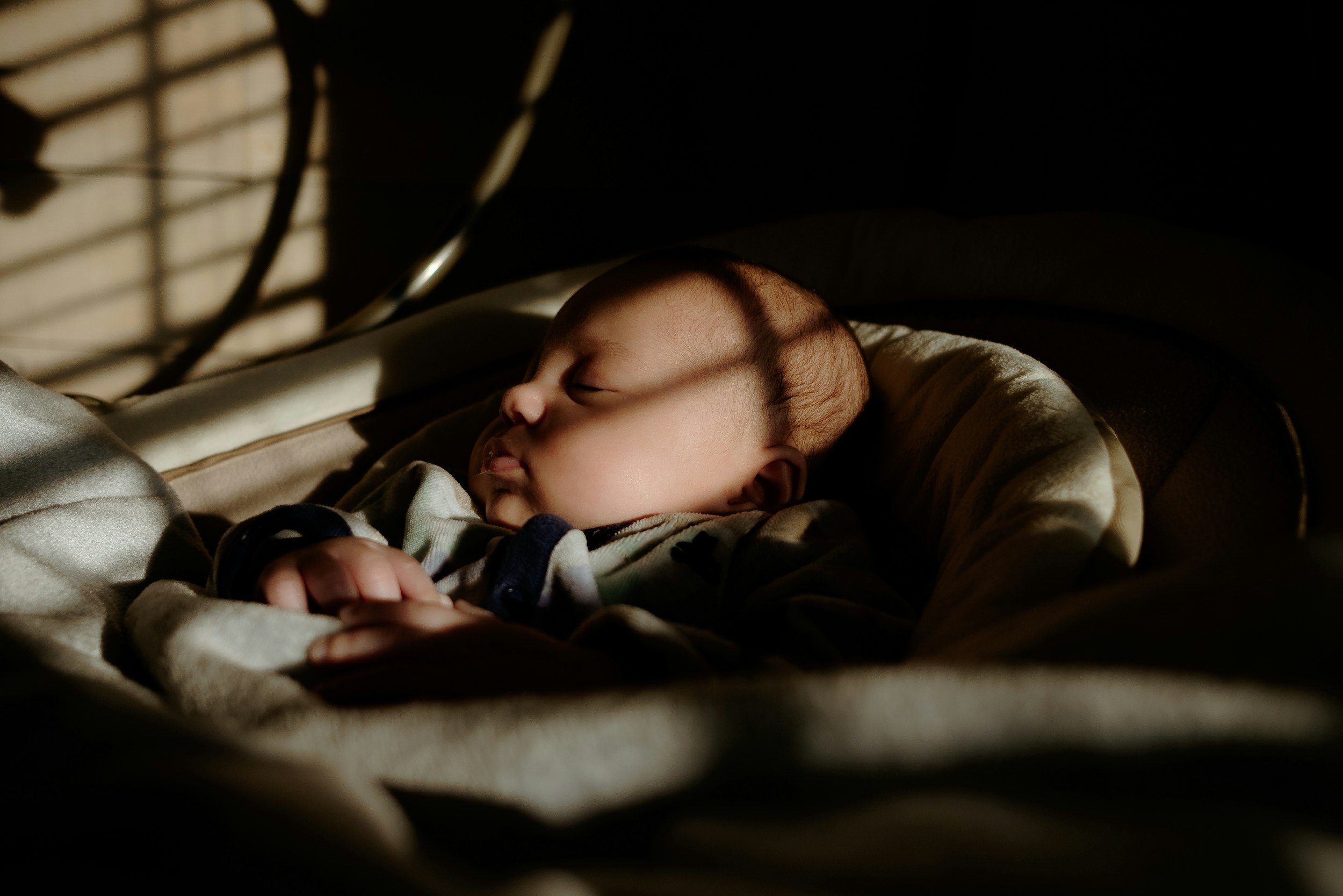 A sleeping baby with light skin, lying in a carrier with sunlight casting shadows across the face, closed eyes, and relaxed facial expression.