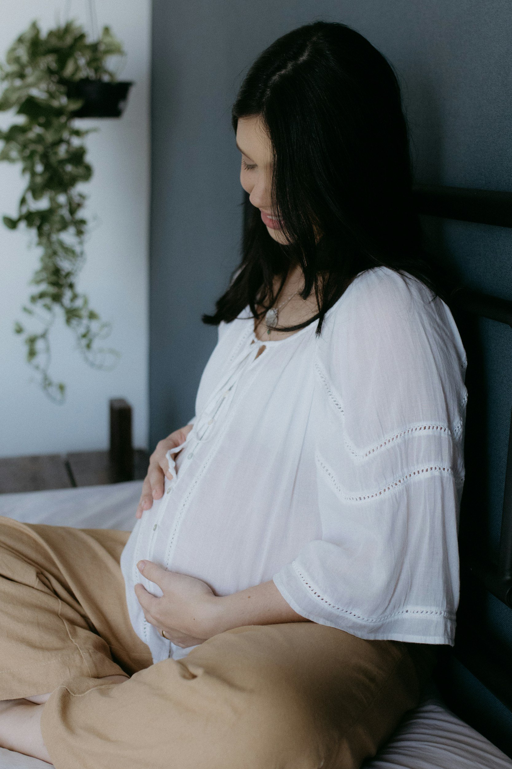 A woman with black hair, wearing a white blouse and beige pants, sitting and holding her pregnant belly with both hands. She is resting against a gray wall, with a plant on a shelf in the background.
