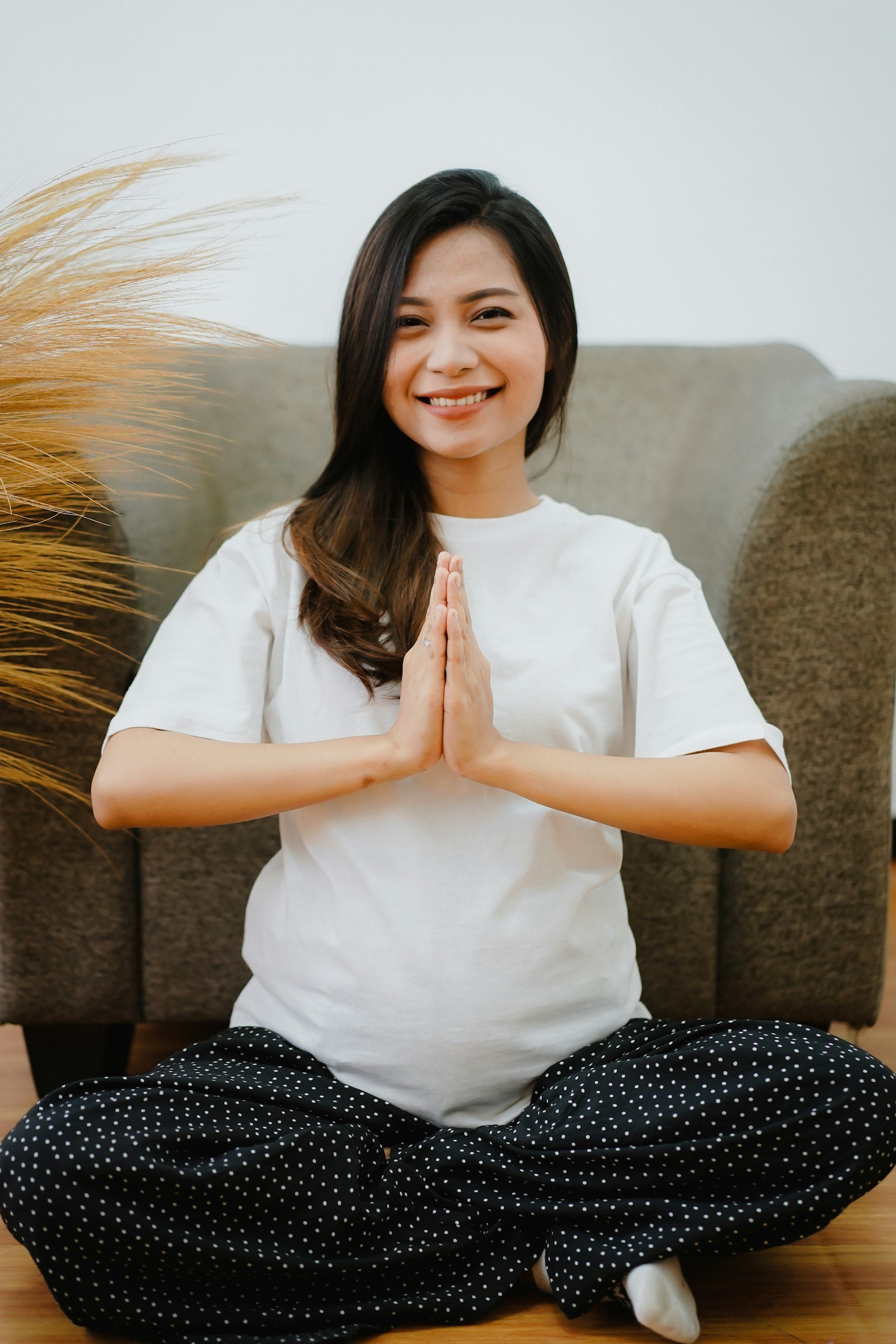 A woman sitting cross-legged on a wooden floor, smiling, with hands pressed together in a prayer position, wearing a white t-shirt and black polka dot pants, in front of a brown cushioned wall and a beige decorative item on the left side.