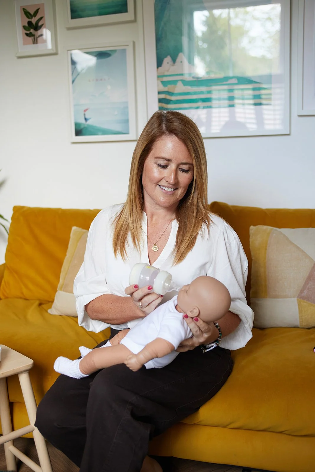 A woman with red hair feeds a baby from a bottle while sitting on a yellow couch in a living room with framed artwork on the wall.