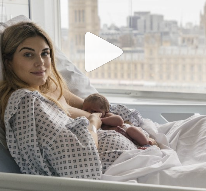 A woman in a hospital bed holding her newborn baby, with city buildings visible through a window in the background.