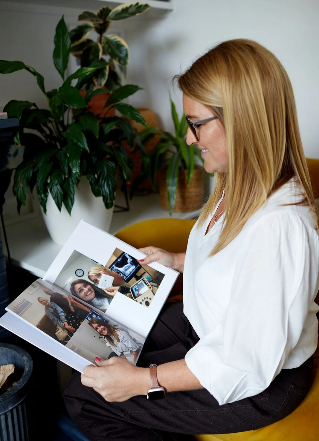 A woman with long blonde hair, glasses, and a white shirt sitting in a yellow chair, looking at a photo album with pictures of herself and others, and smiling. There are green plants and a clock on the wall in the background.