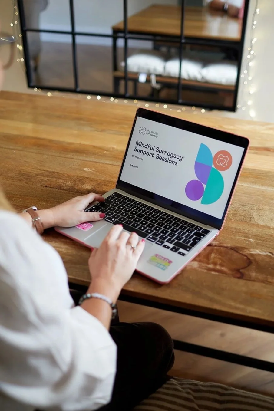 Woman using a laptop on a wooden table, screen displaying 'Mindful Surrogacy Support Sessions'.