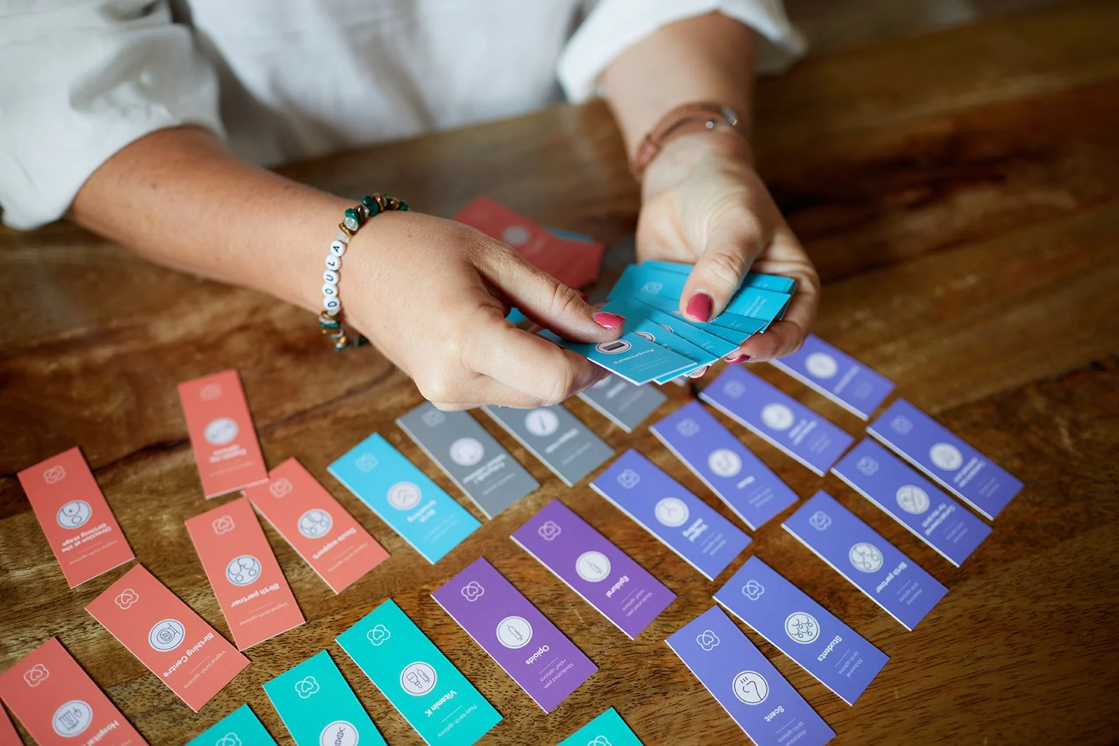 A person with pink nail polish and a beaded bracelet is holding a card from a set of colorful cards with icons and text, seated at a wooden table with more cards laid out.