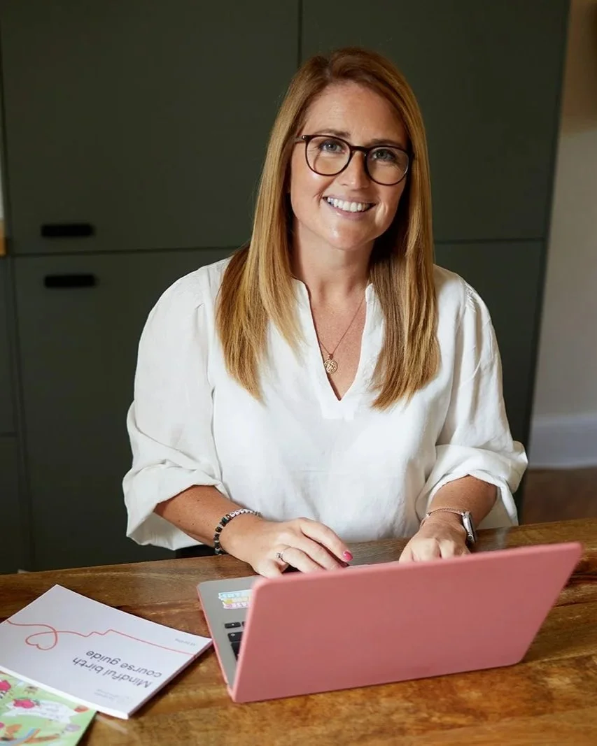 Emiliana Hall wearing glasses, and a white blouse sitting at a wooden table using a pink laptop.
