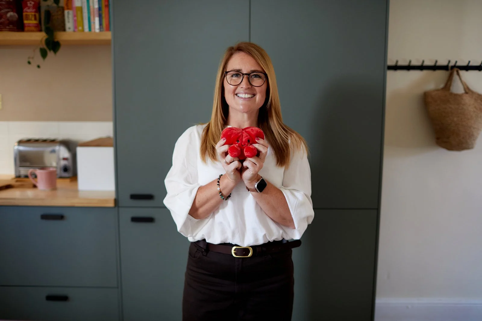 Emiliana Hall smiling and holding a red plush teddy bear in a kitchen. She is wearing a white blouse, a black skirt, and a smartwatch. The kitchen has blue cabinets, a wooden countertop, a toaster, a pink mug, a bookshelf.