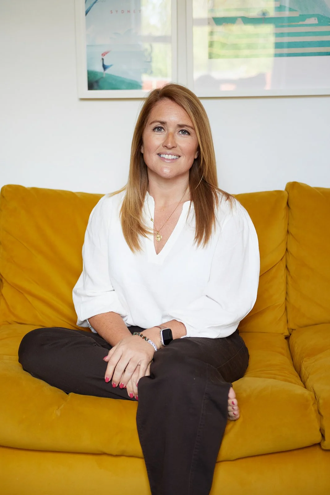 Emiliana Hall wit sitting cross-legged on a yellow couch, smiling at the camera, wearing a white blouse, black pants, and a smartwatch.