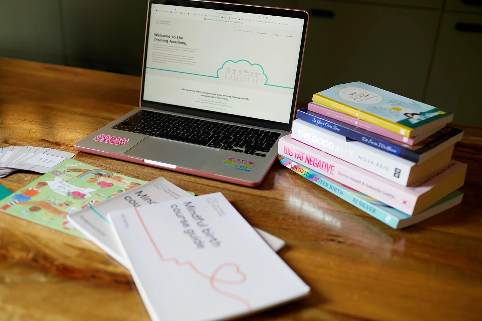 A wooden desk displaying a laptop, a stack of books, some pamphlets, and colorful brochures.