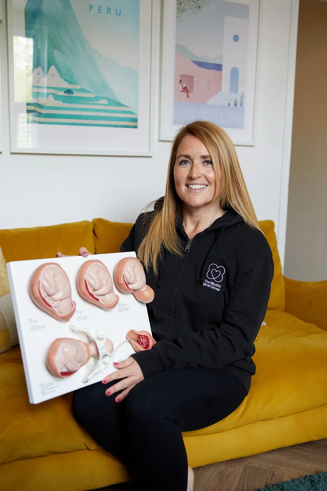 A woman with long red hair sitting on a yellow couch, holding a diagram of the female reproductive system. She is wearing a black hoodie with a logo and text on it. Behind her, there are two framed pictures on the wall, and the room has a cozy and modern ambiance.