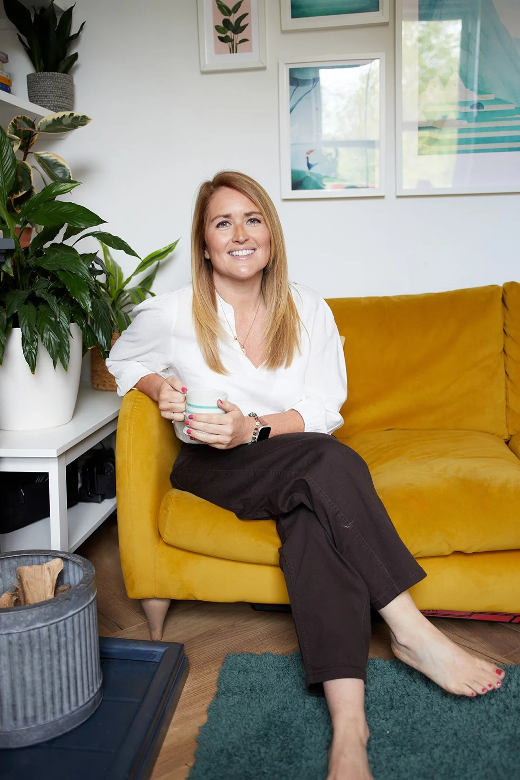 Emiliana Hall sitting on a mustard yellow sofa, holding a mug, in a living room with potted plants and framed art on the wall.