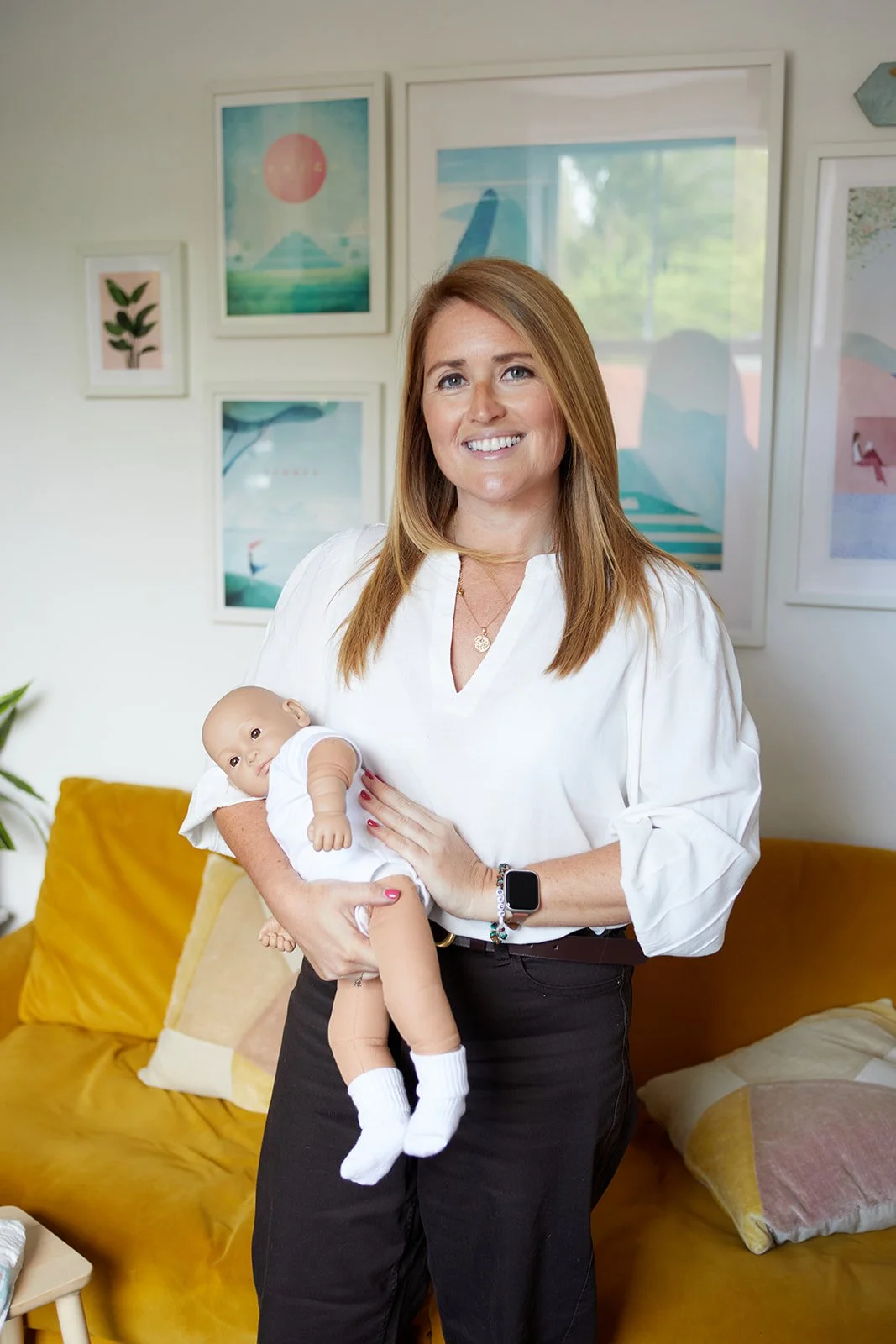 Emiliana Hall smiling while holding a baby in her arms, in a brightly decorated room with framed artwork on the wall and a yellow couch.