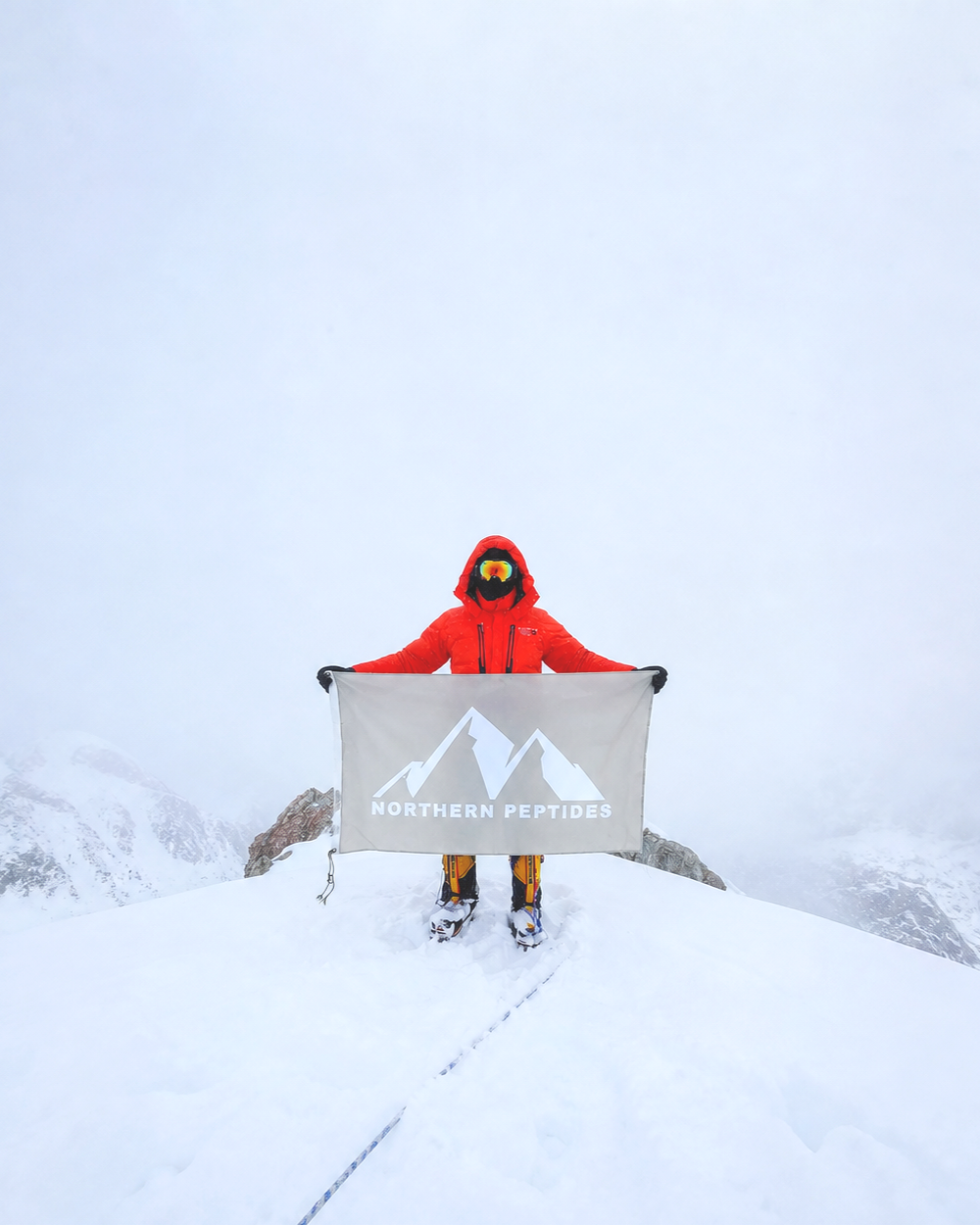 Person in red winter gear holding a Northern Peptides banner on snowy mountain summit amid foggy conditions.