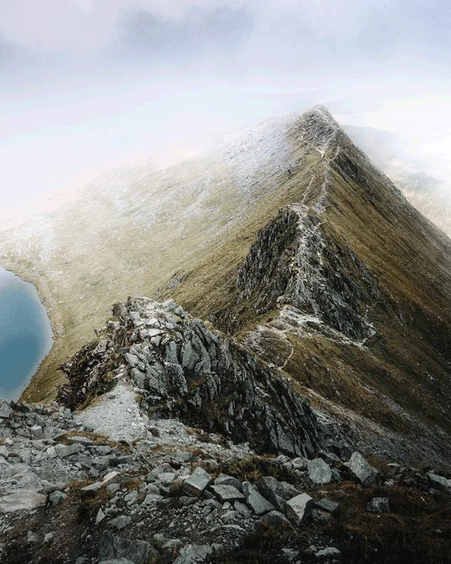 A rugged mountain ridge with a narrow trail, surrounded by rocky terrain and a small lake in the lower left corner, with foggy weather.