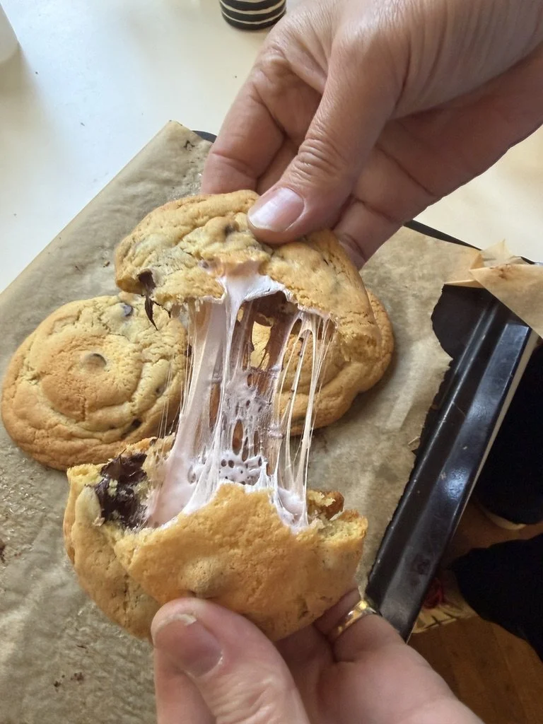 Person pulling apart a large chocolate chip cookie with melted chocolate and gooey melted caramel inside, on a parchment-lined tray
