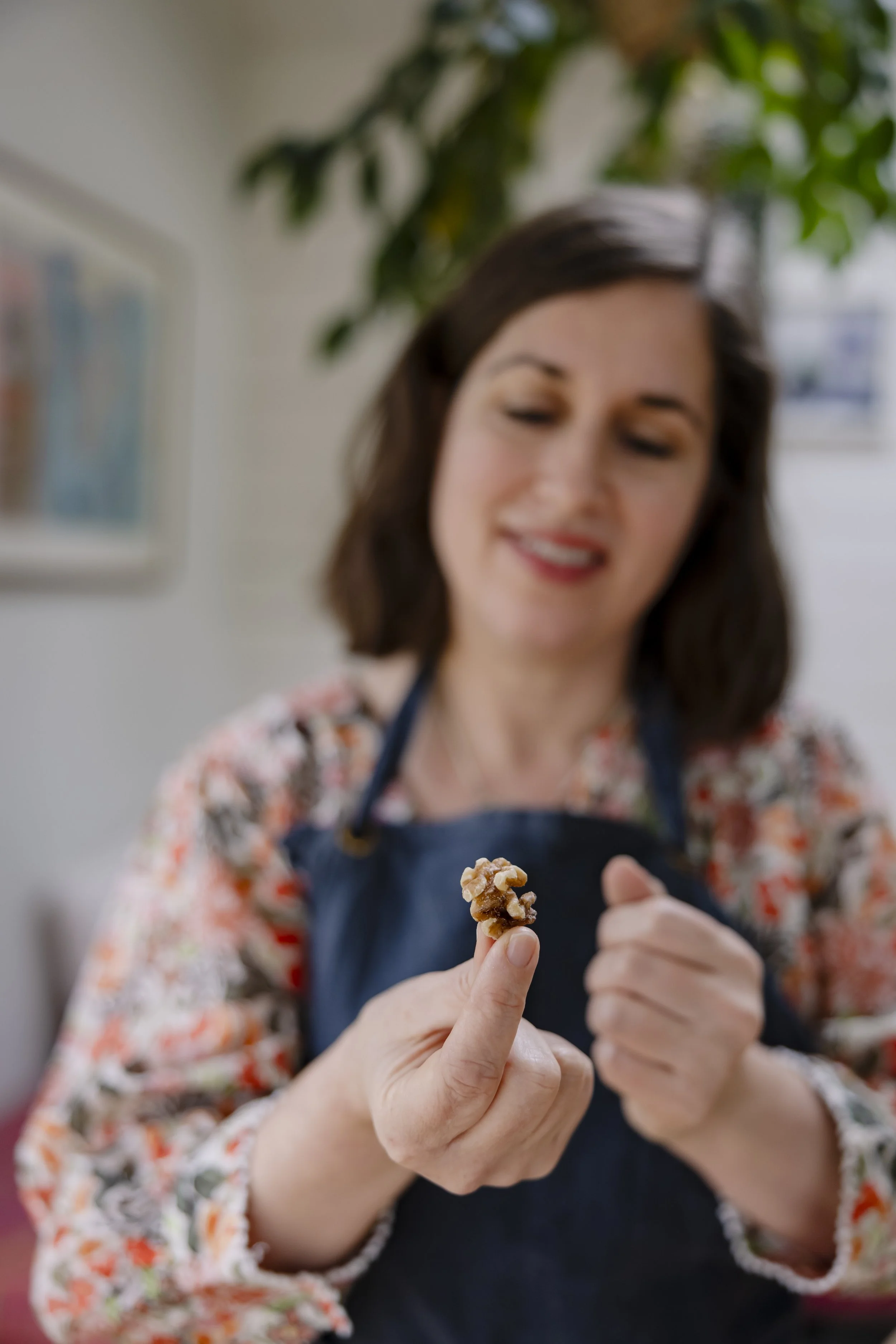 A woman with dark hair, smiling and looking down, holding a piece of food while wearing an apron, with blurred background of houseplants and furniture.