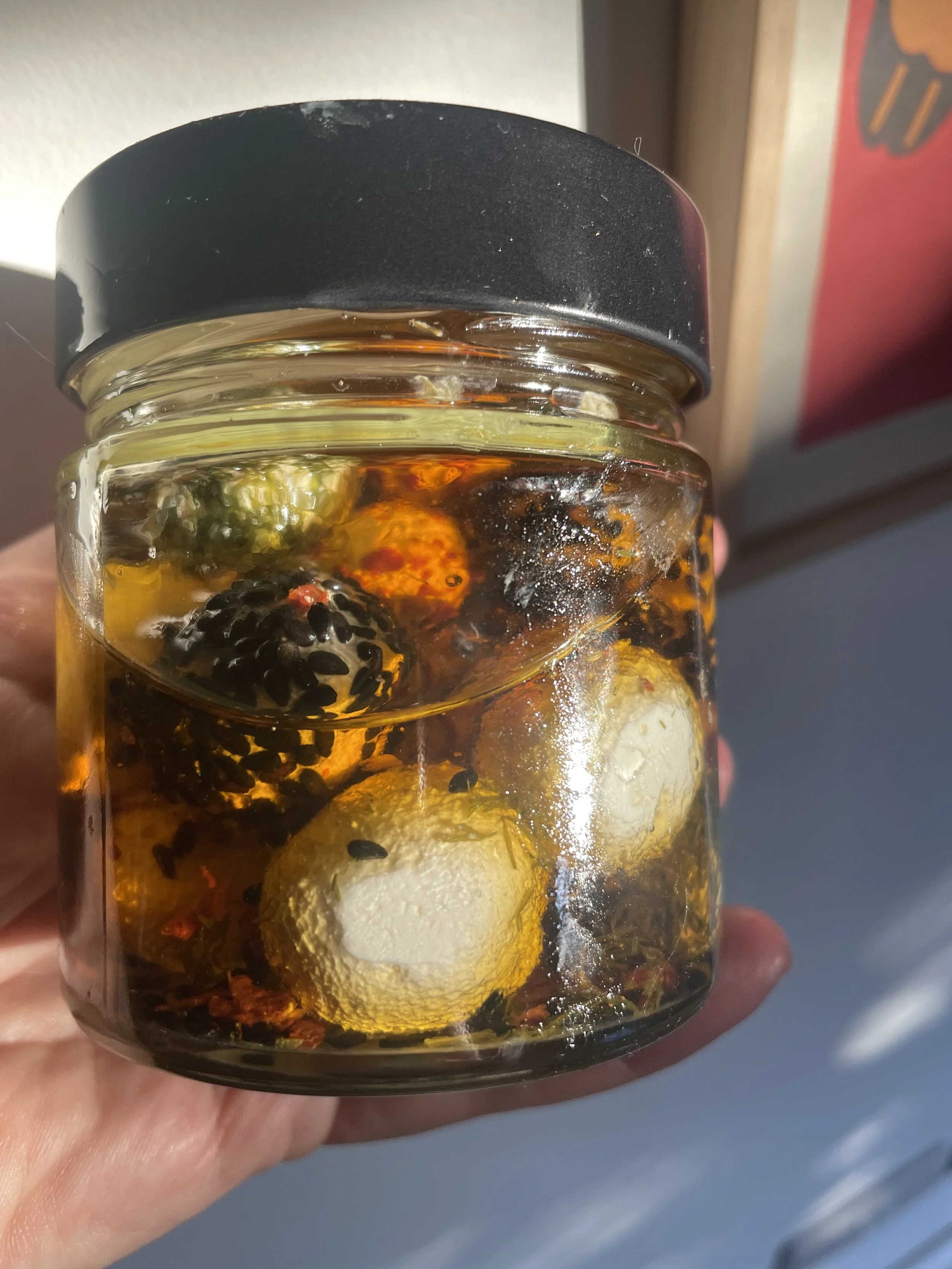 A small glass jar filled with honey, containing assorted herbs and herbs, held in a person's hand with a blurred background.