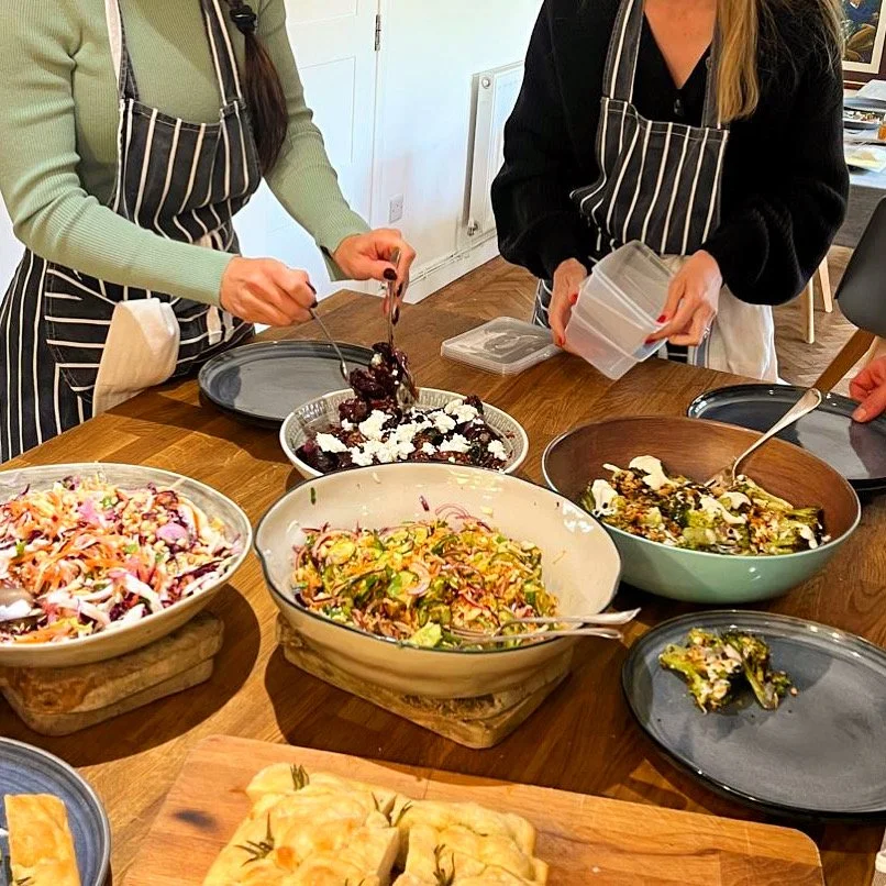 Two women wearing striped aprons preparing a table with various dishes, including salads and desserts, in a dining room.