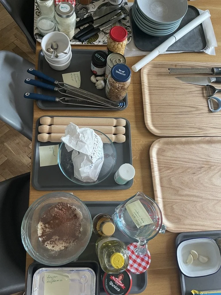 Organized kitchen preparation for baking, with various baking ingredients, utensils, and tools on black trays and wooden cutting boards.