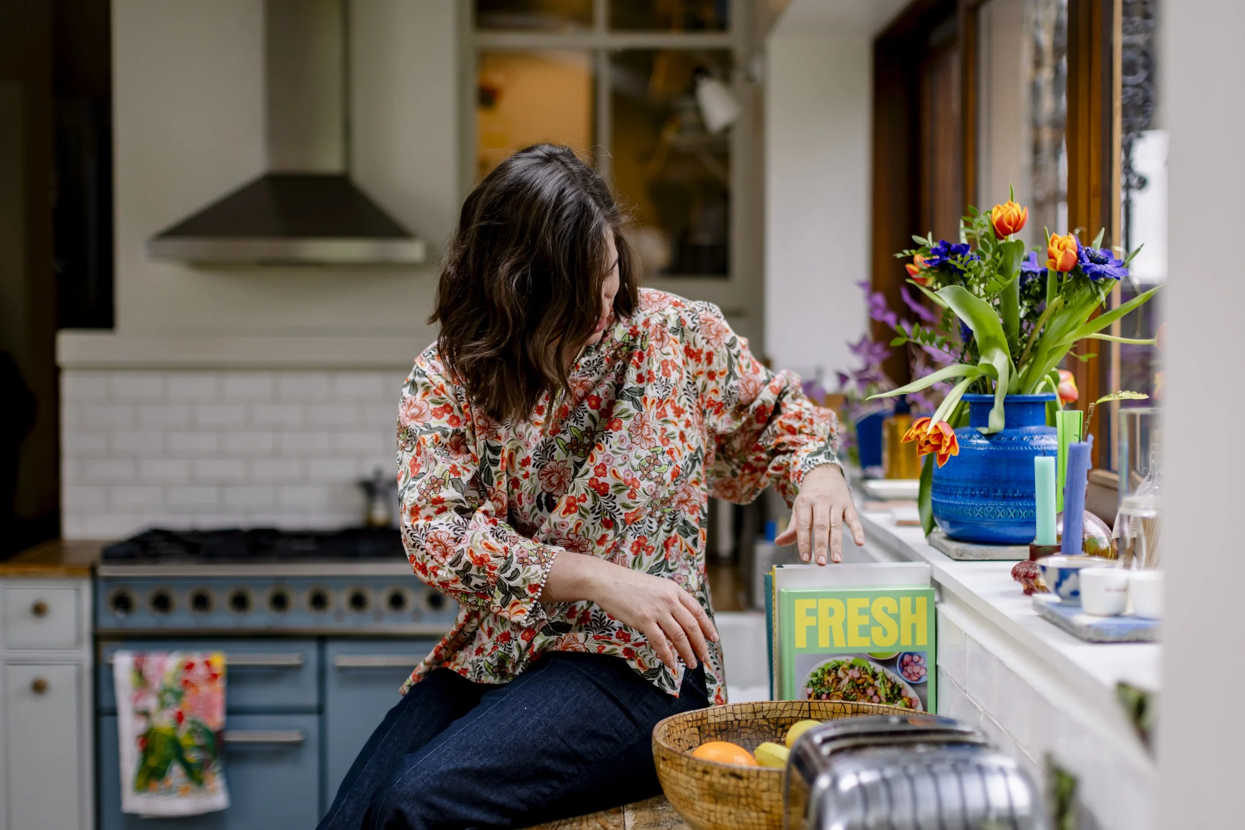 , curly hair wearing a floral blouse in a kitchen. She is sitting at a counter, possibly preparing food. There is a large blue vase with colorful flowers including tulips and other blooms on the counter. A sign with the word "FRESH" and pictures of food is also visible on the counter.