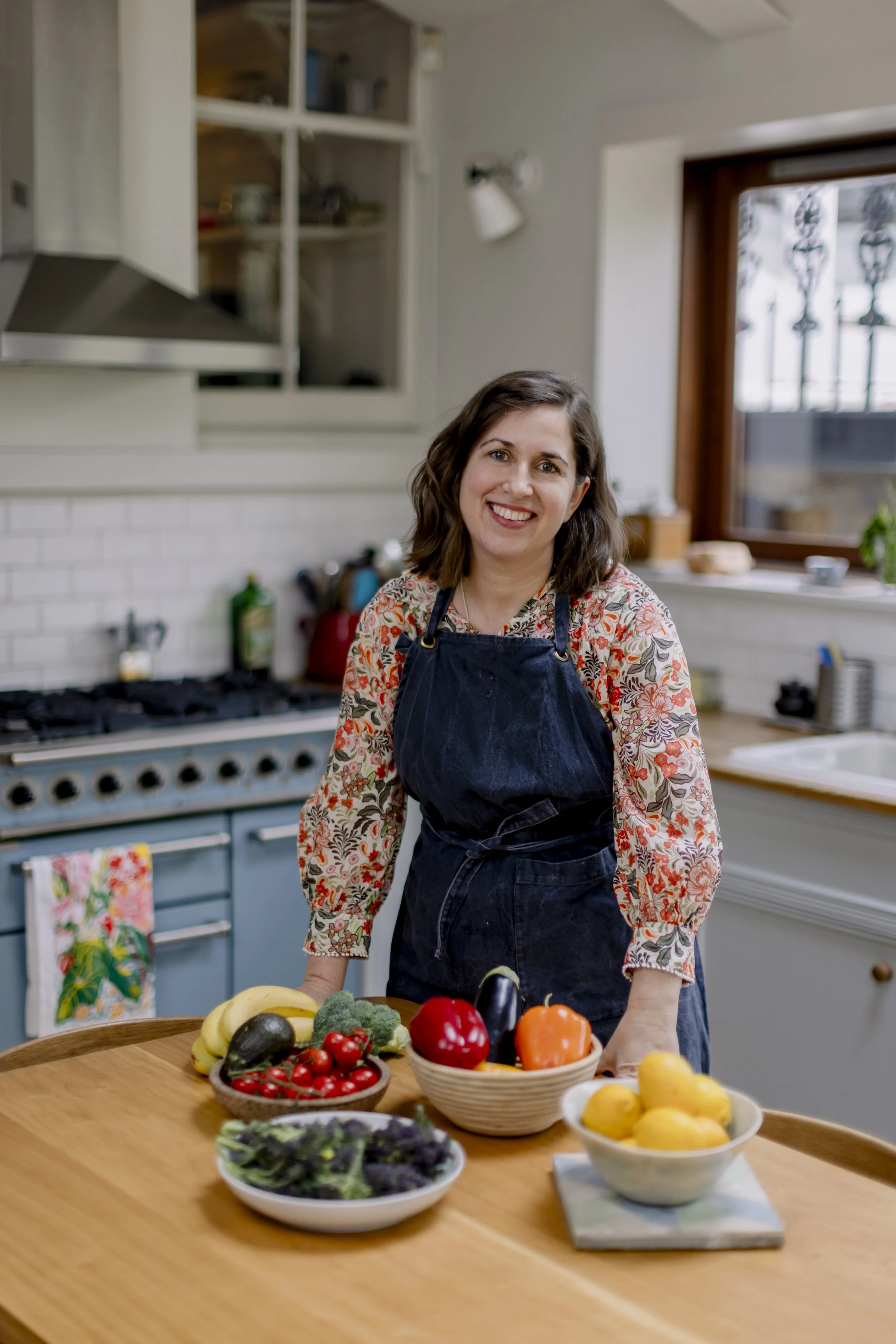 A woman with shoulder-length brown hair, wearing a floral shirt and a dark apron, standing in a kitchen behind a wooden table with bowls of fresh vegetables and fruits, smiling at the camera, with a window and kitchen cabinets in the background.