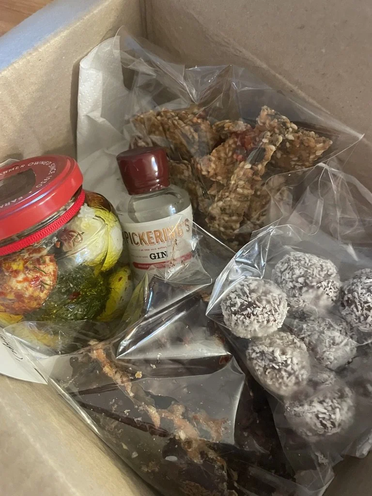 Assorted homemade treats including chocolate-covered balls coated with shredded coconut, granola bars, pickled vegetables in a jar, and a small bottle of gin inside a cardboard box.