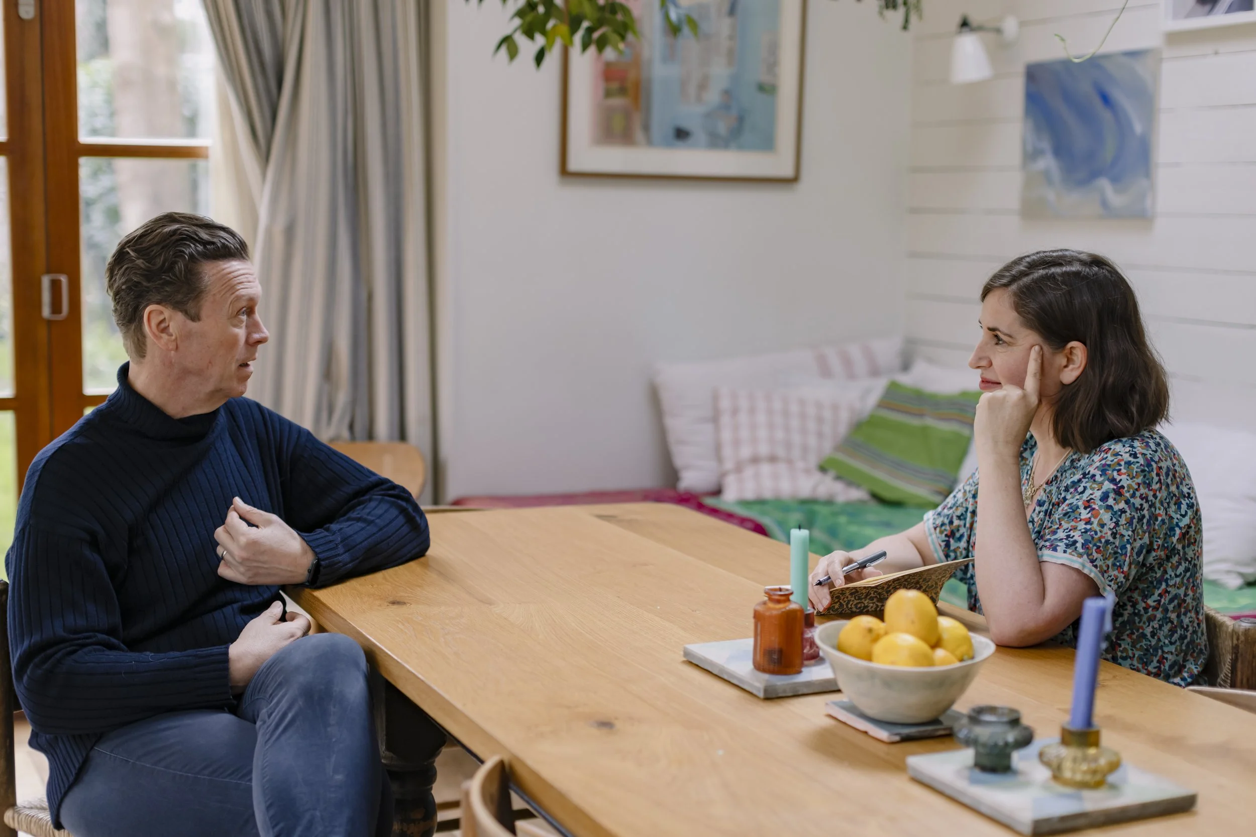 A man and a woman sitting at a wooden dining table having a conversation. The man is wearing a dark blue sweater, and the woman is wearing a colorful patterned shirt. There are kitchen items and fruit on the table, with a window, artwork, and a bed in the background.