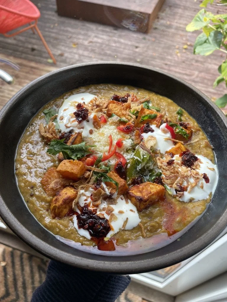 A bowl of Indian-style curry topped with yogurt, coriander, red chili slices, and dried herbs, with chunks of vegetables and proteins visible in the sauce.