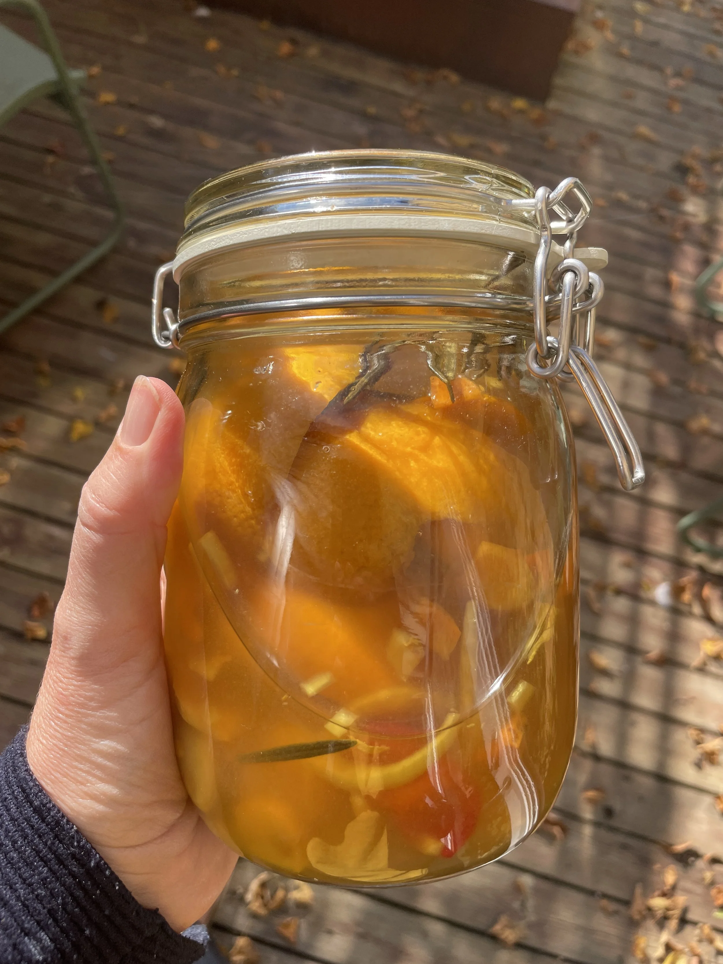 A close-up of a hand holding a glass jar filled with yellow-orange liquid and slices of fruit, outdoors on a wooden deck with fallen autumn leaves.