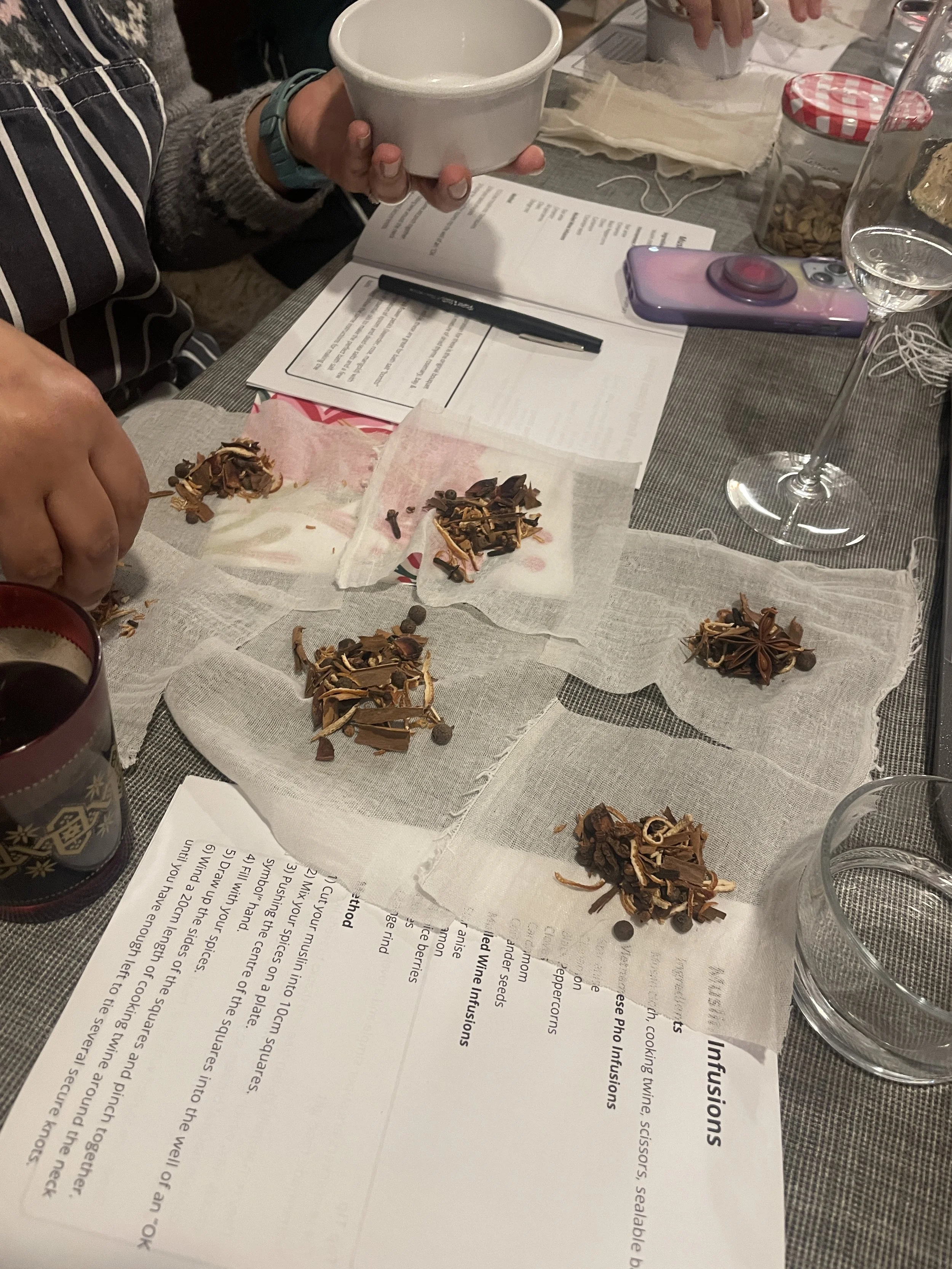 Table set with sheets of paper, a black pen, a clear glass of water, a purple camera, various jars, and dried spices, with a person's hand holding a white bowl.