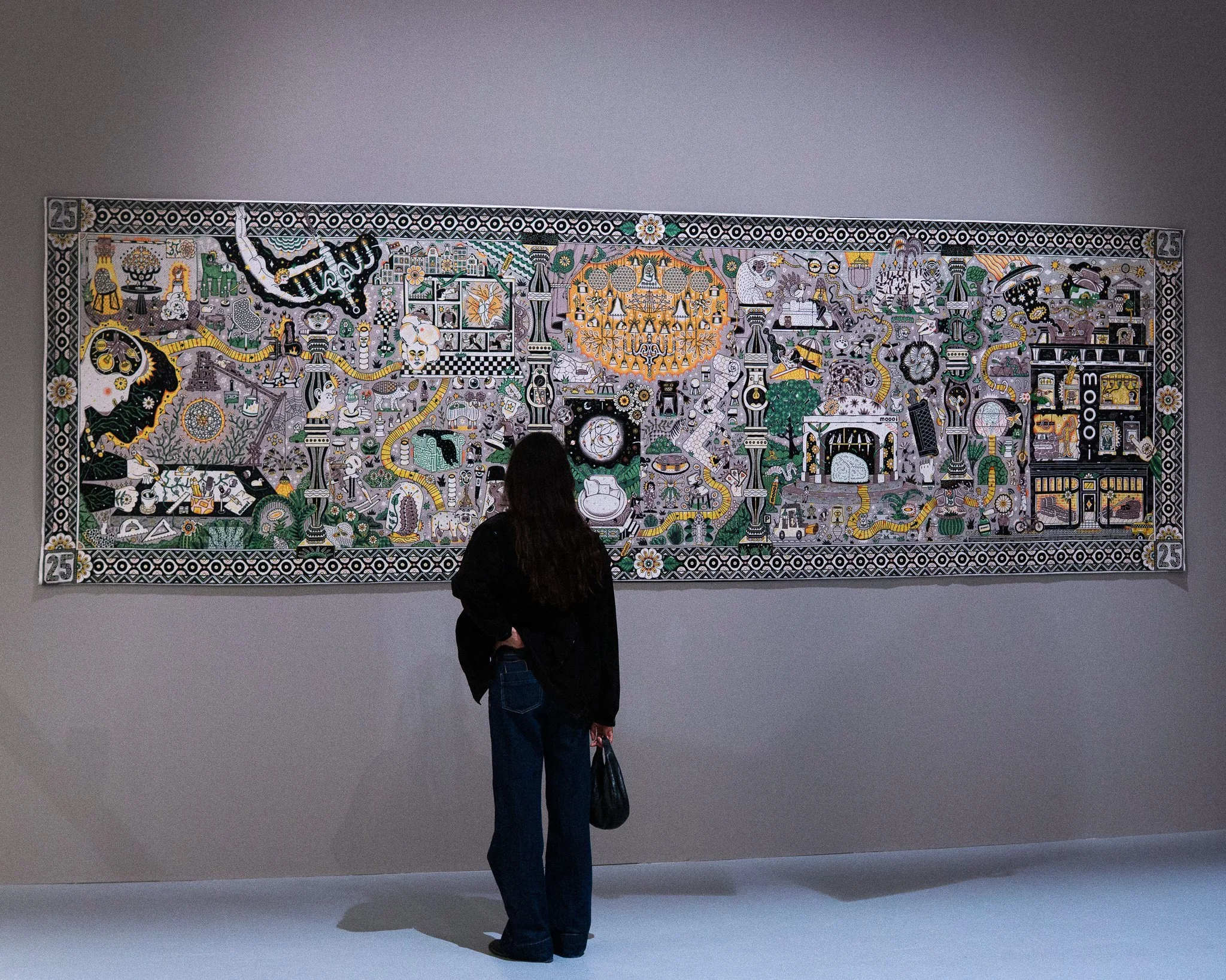 Person with dark hair wearing a black jacket and blue jeans standing in front of a large, detailed artwork in a museum or art gallery.  Tapestry and jacquard weaving art