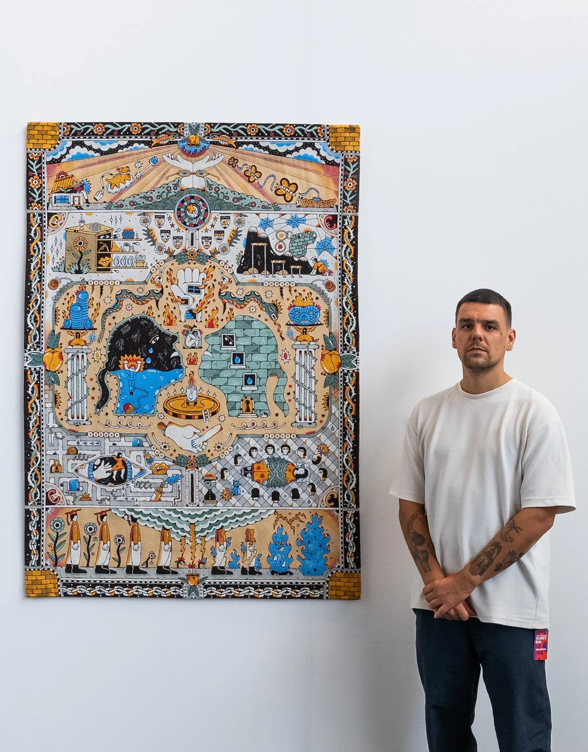 A man with tattoos on his arms stands next to a colorful, abstract artwork on a white wall in an art gallery. Tapestry and jacquard weaving art