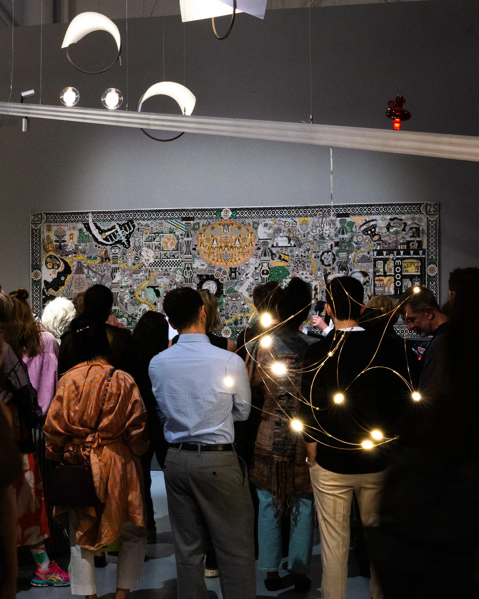 People standing inside a room with a large, colorful, detailed mural on the wall behind them, with some wearing casual clothing and some with hats or glasses, and one person in the foreground is wearing a striped orange and beige outfit. 