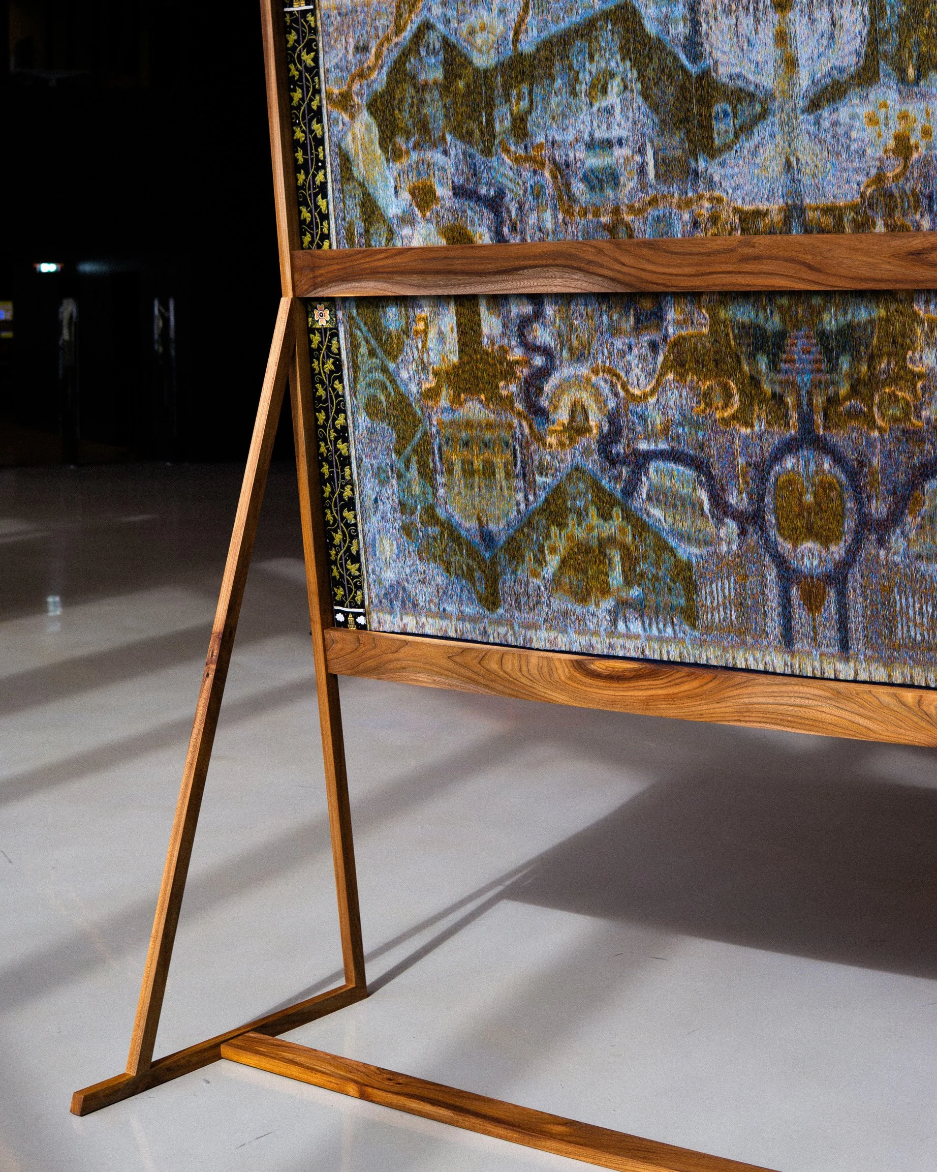 A wooden easel displaying a tapestry or rug with an intricate colorful landscape design, featuring trees, water, and mountains, in an art gallery or exhibition setting.  Tapestry and jacquard weaving art