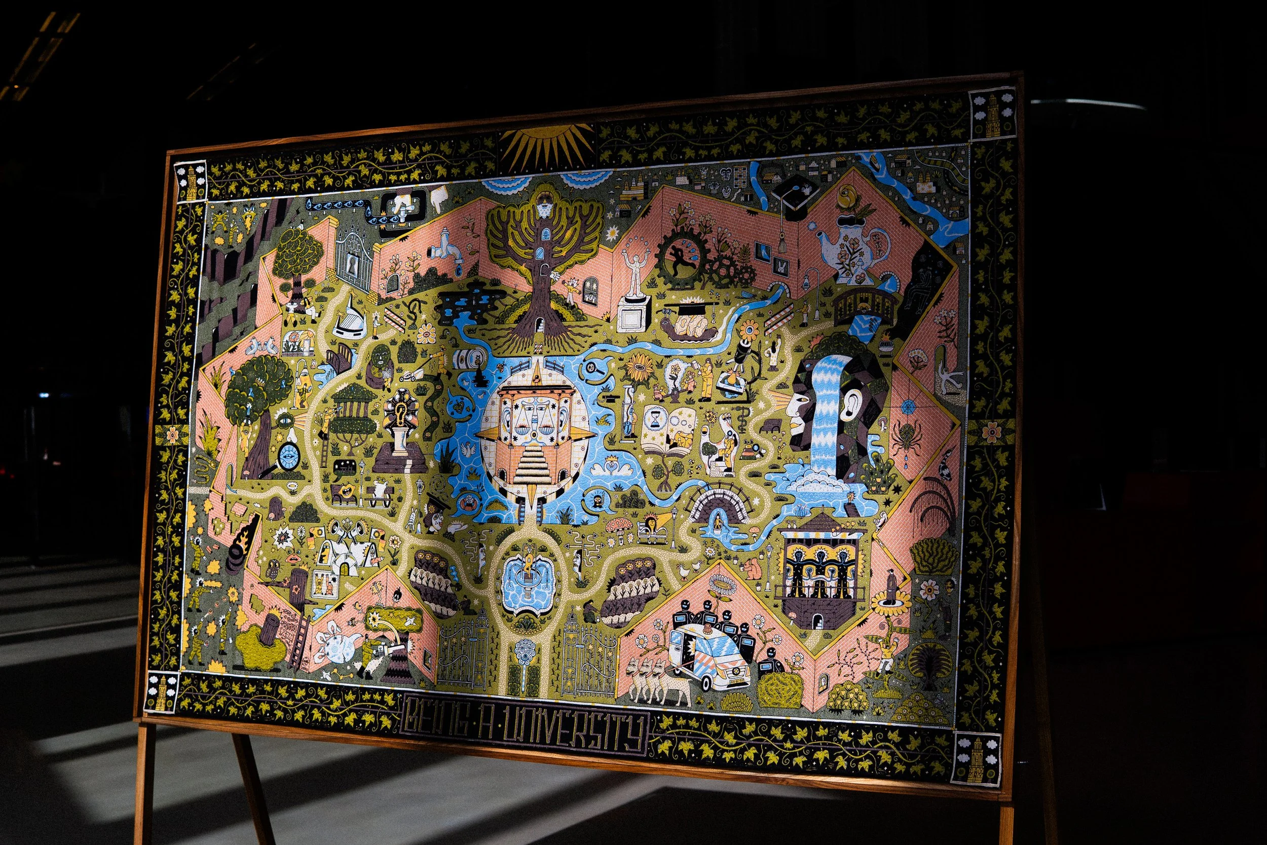 Colorful artwork on a framed canvas depicting a garden scene with various trees, fountains, animals, human figures, and abstract elements, titled 'Being a University'.  Tapestry and jacquard weaving art